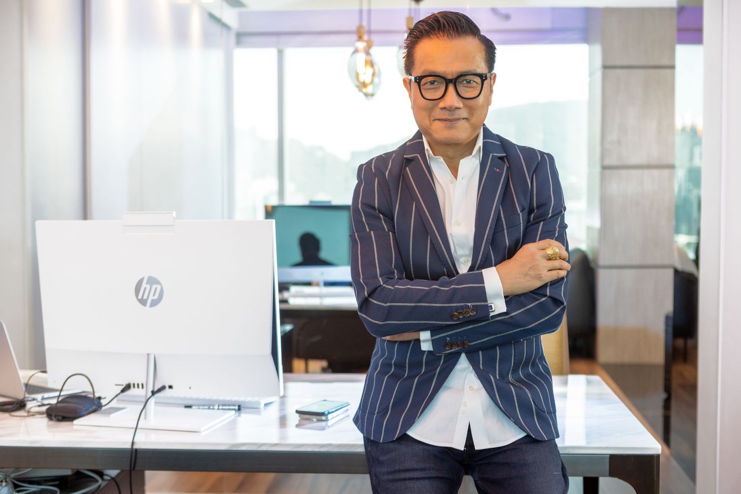Professional in pinstriped navy suit with glasses stands confidently in modern office with laptop on desk.