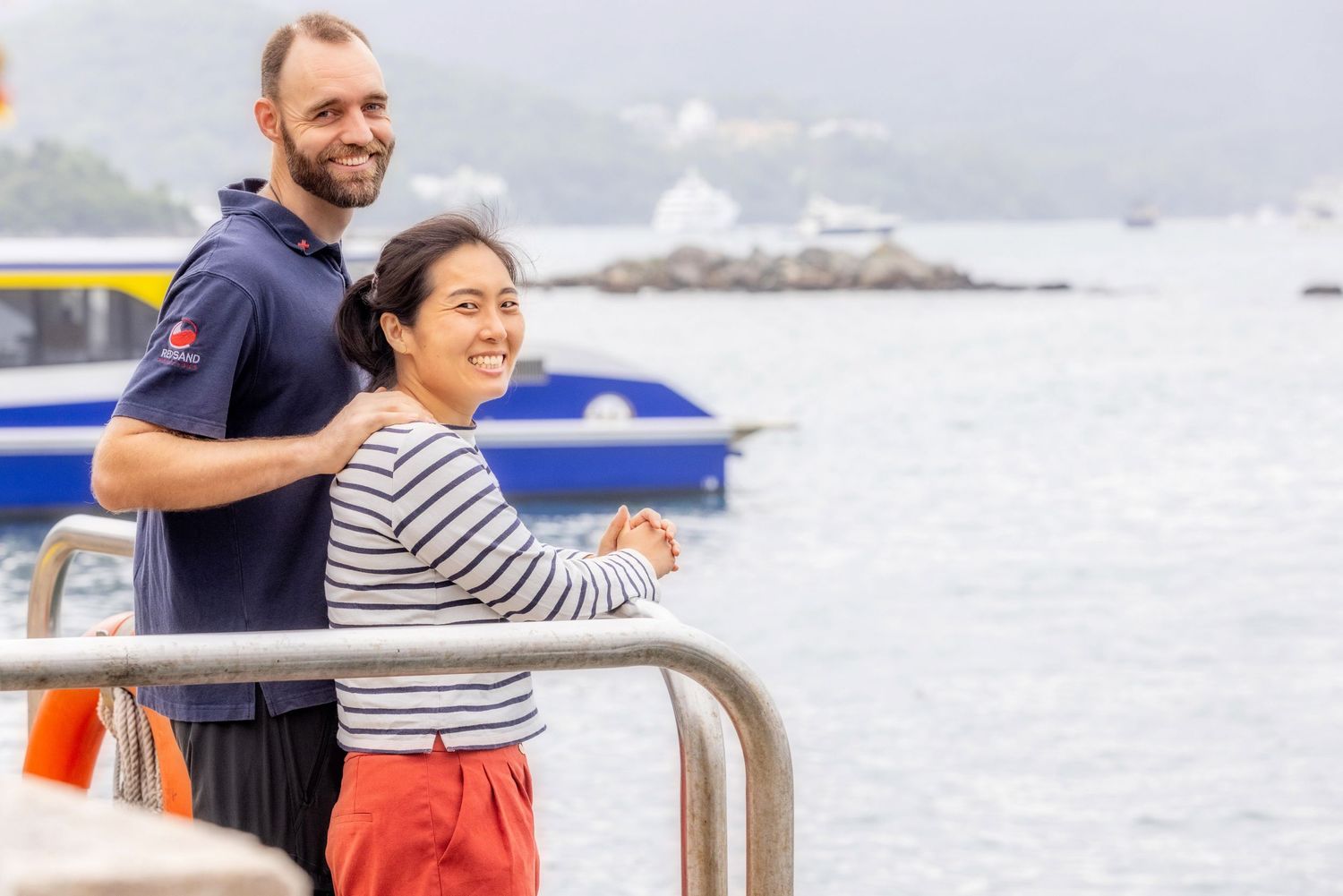 Two people enjoying the view from a boat railing on a misty day near a lighthouse and coastline.