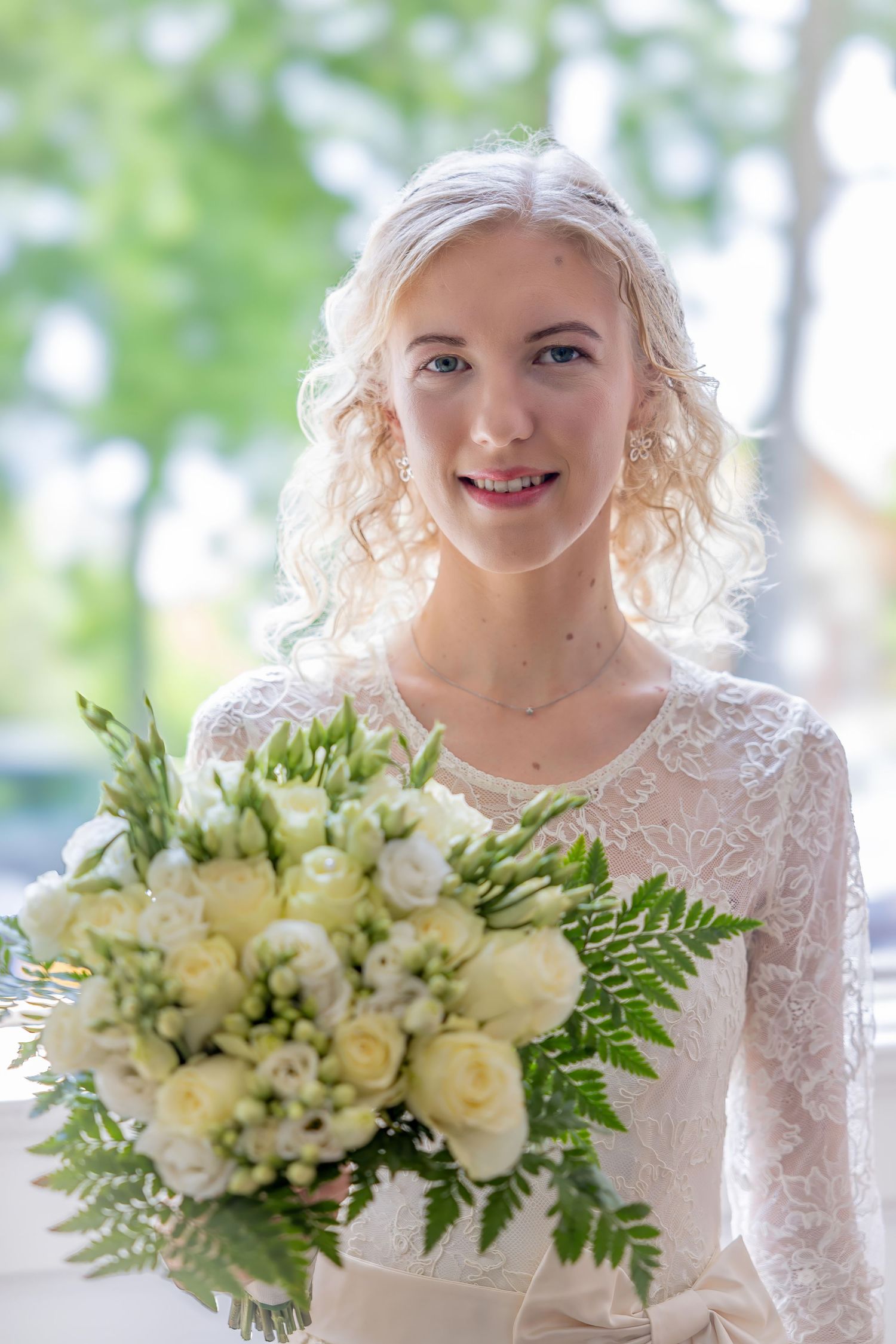A bride in a white lace dress holds an elegant bouquet of cream roses and ferns on her wedding day.