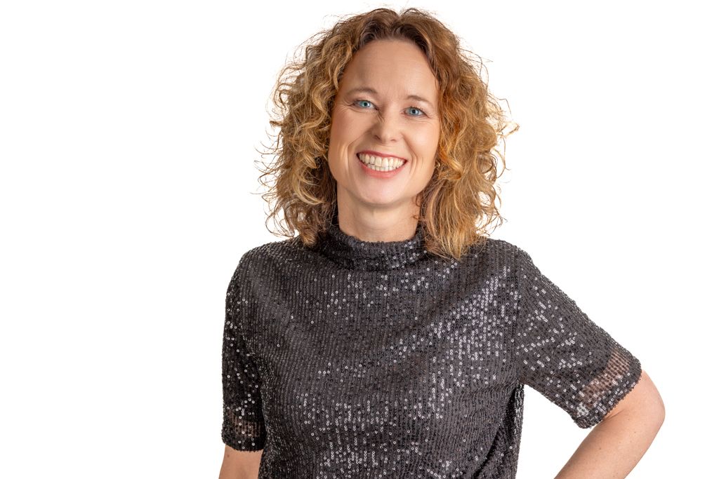 A woman with curly red hair wearing a sparkly black top poses with her hand on her hip against a white background.