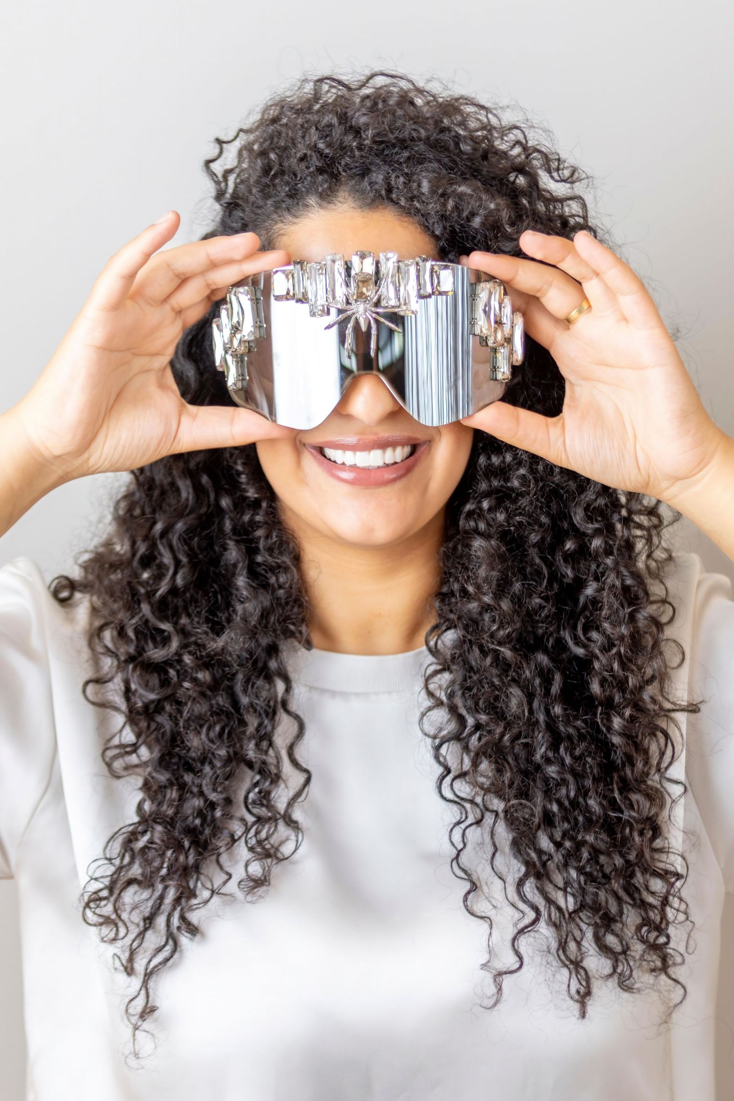Person with long curly hair holds goggles in front of their eyes while smiling in a bright setting.