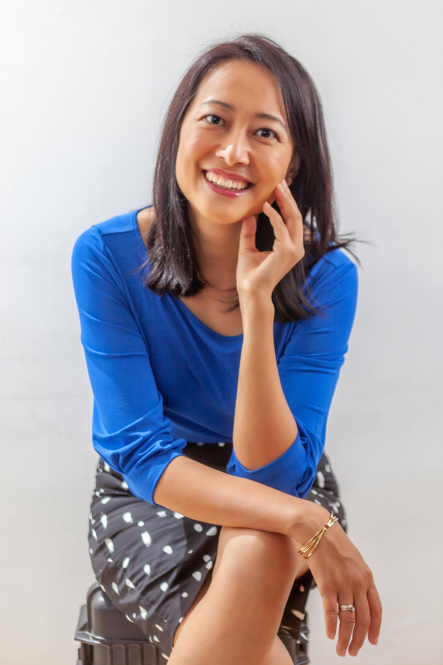 A person in a bright blue top and patterned skirt sits casually while smiling against a light background.