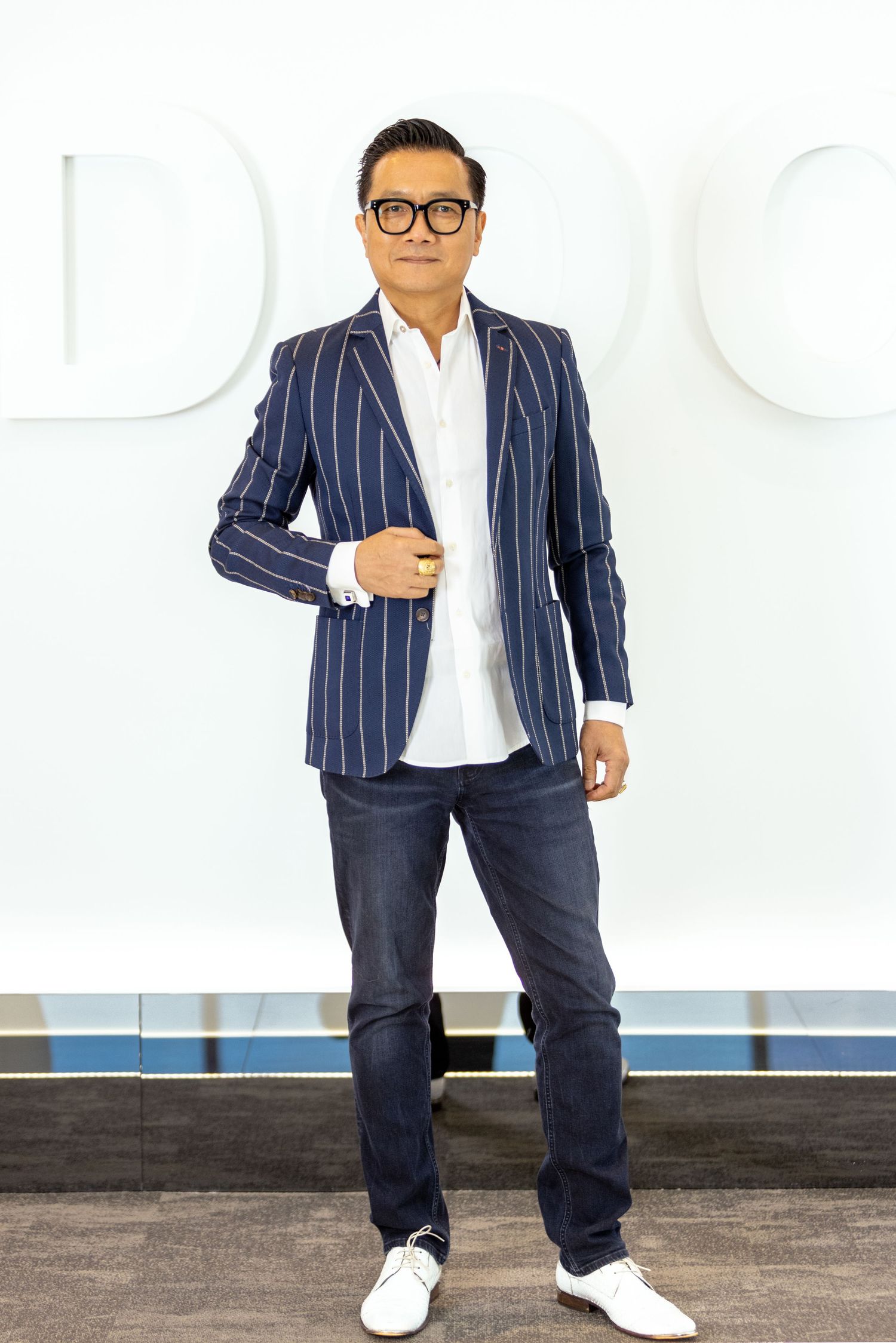 A professional Chinese man in a pinstripe navy blazer, white shirt, dark jeans and white sneakers poses against a white wall.