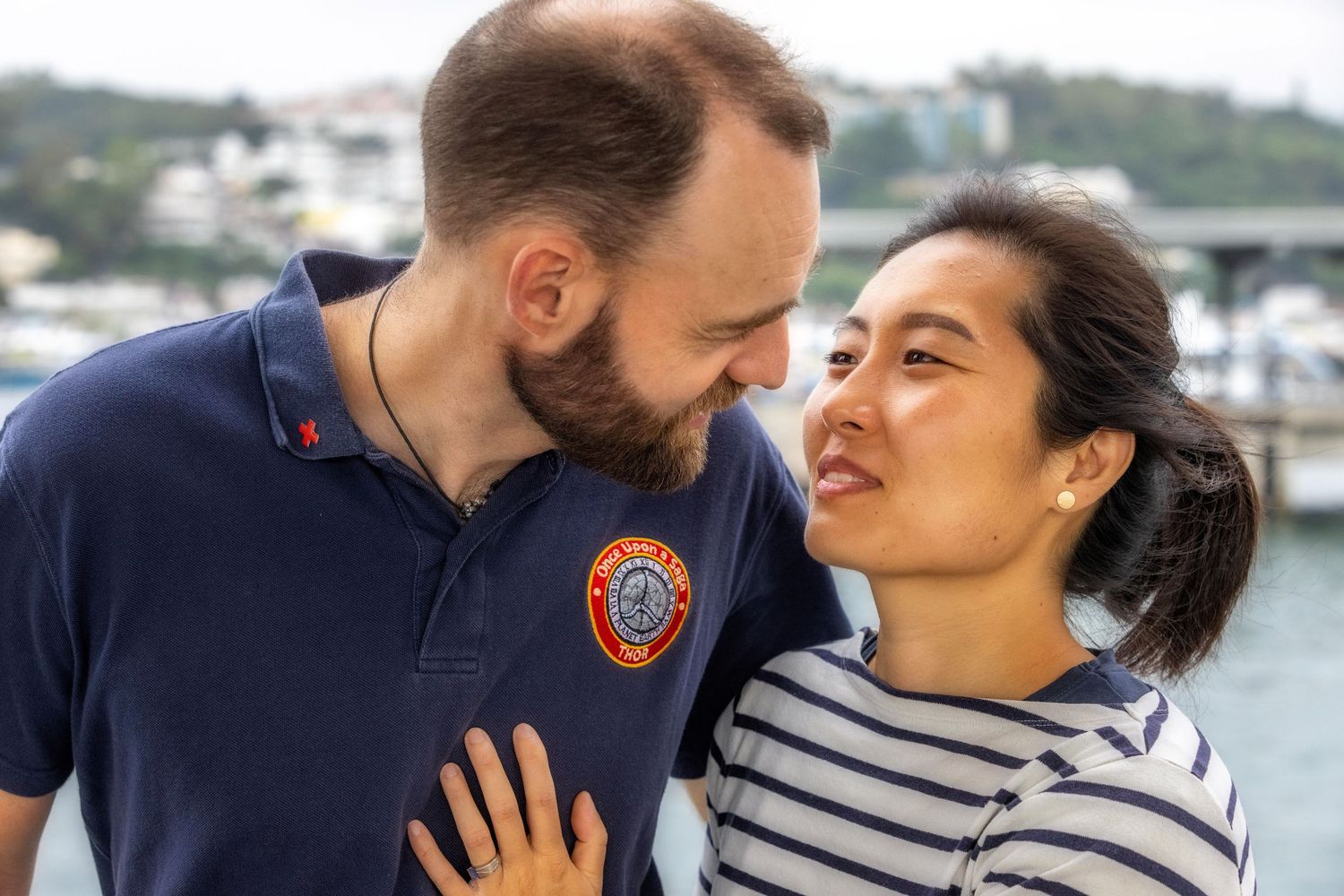 A romantic couple shares an intimate moment by the waterfront, with one wearing a navy polo and the other in stripes.