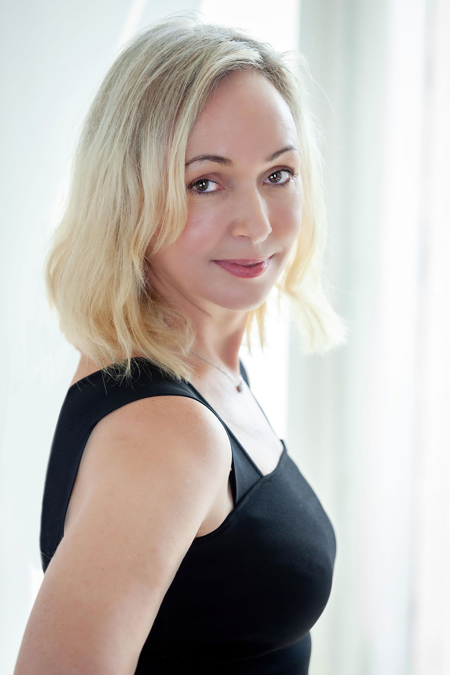 Blonde woman in a black sleeveless top poses with a soft smile against a bright window background.