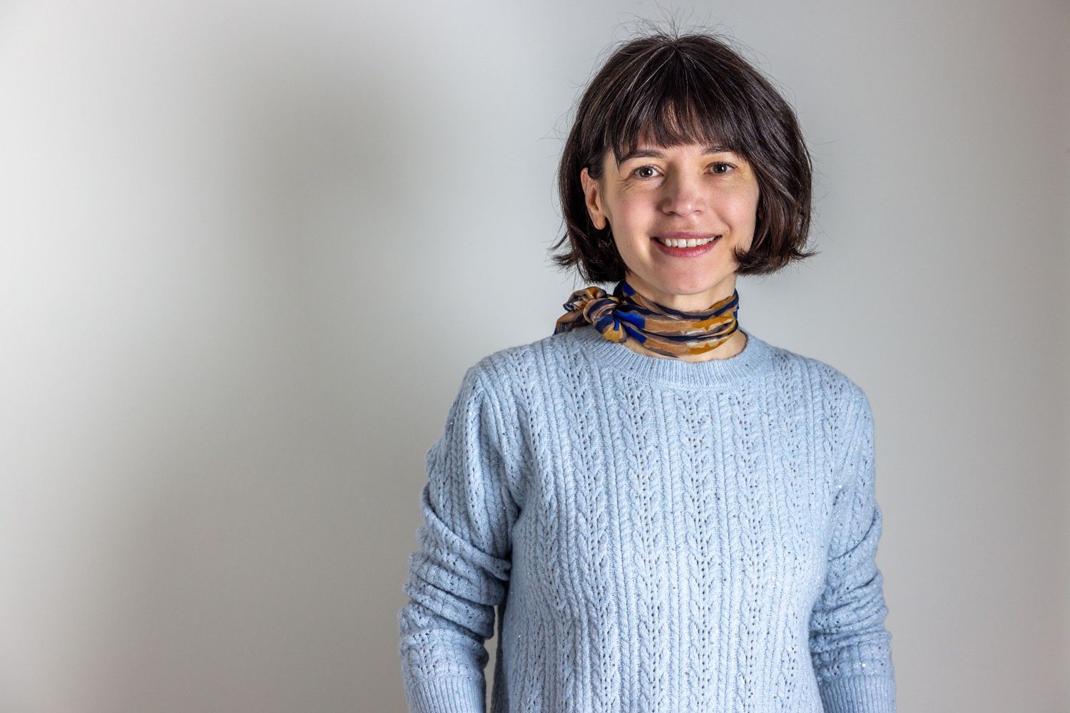 Person wearing a light blue cable knit sweater and scarf smiles against a neutral background.