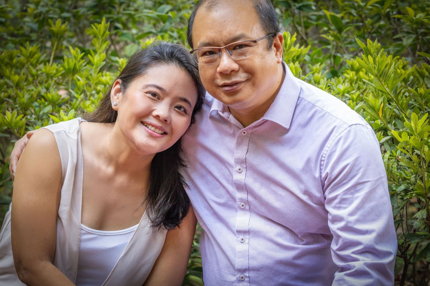 A couple poses close together outdoors with greenery in the background on a sunny day.