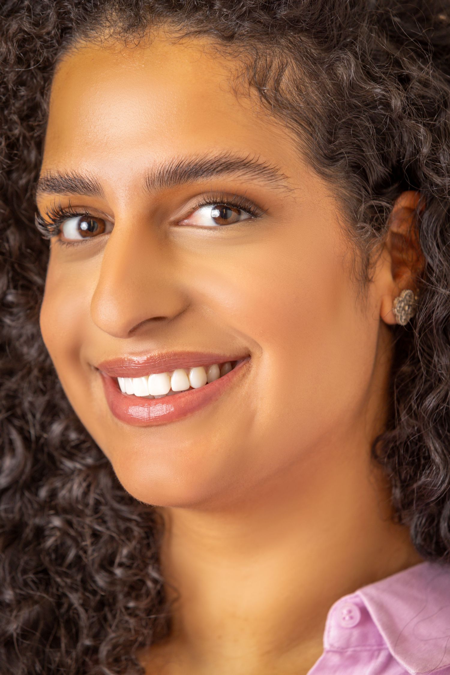 Close up portrait of someone with curly dark hair smiling warmly while wearing a pink shirt.