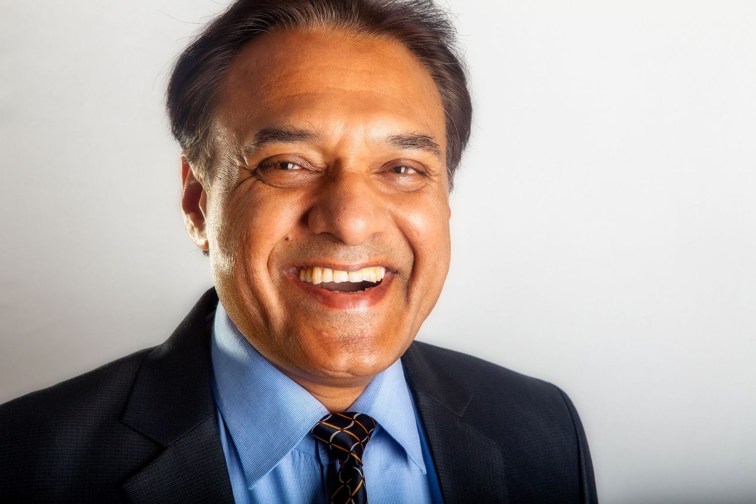 Professional portrait of Indian man in navy suit and blue tie smiling warmly against light background.
