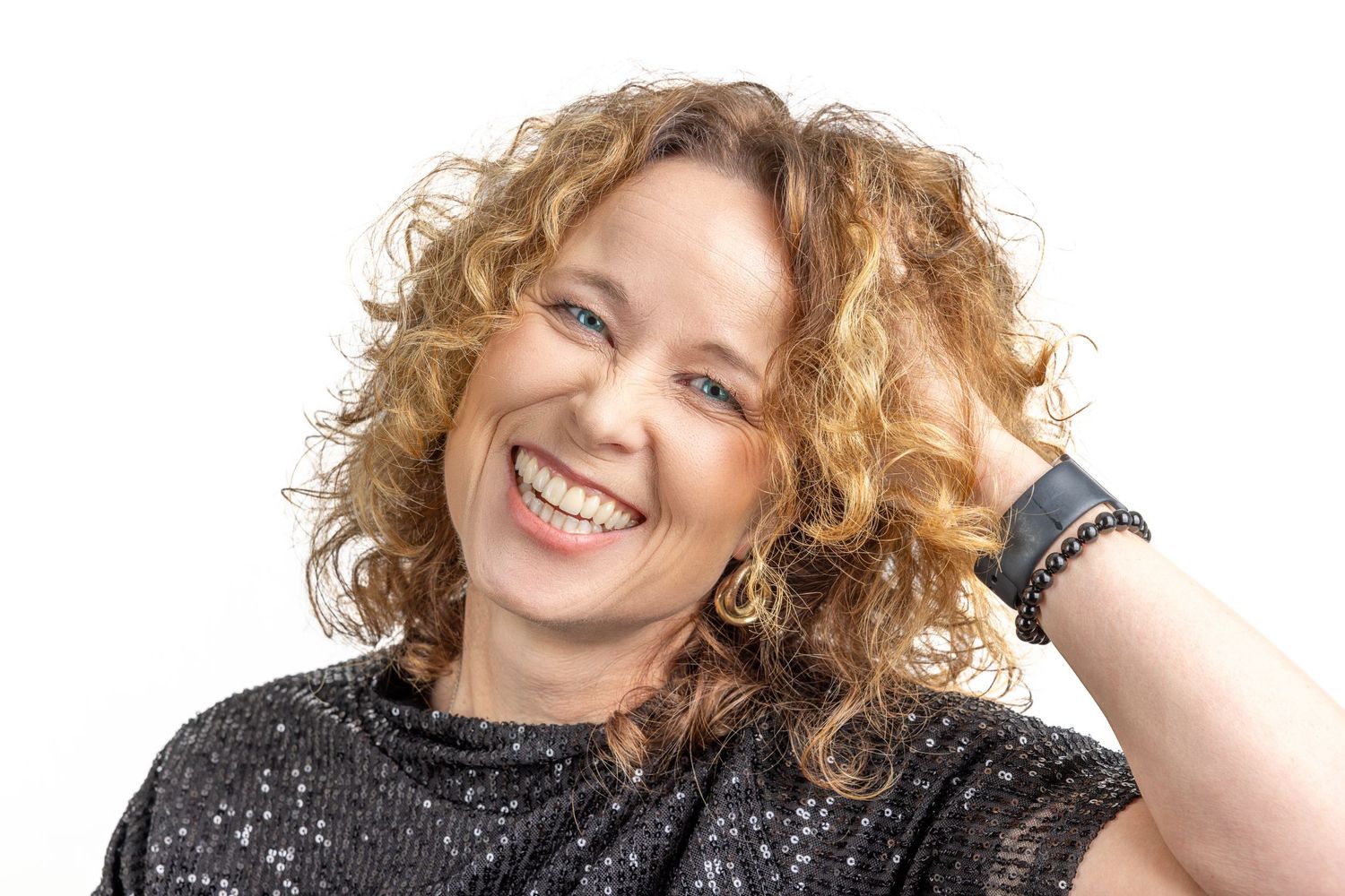A person with curly blonde hair wearing a sequined black top smiles brightly against a white background.