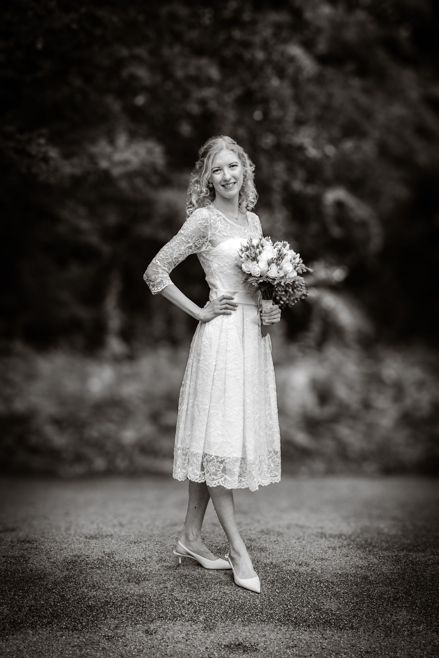 Elegant bride in vintage-style tea length lace dress poses outdoors with bouquet in black and white photography.