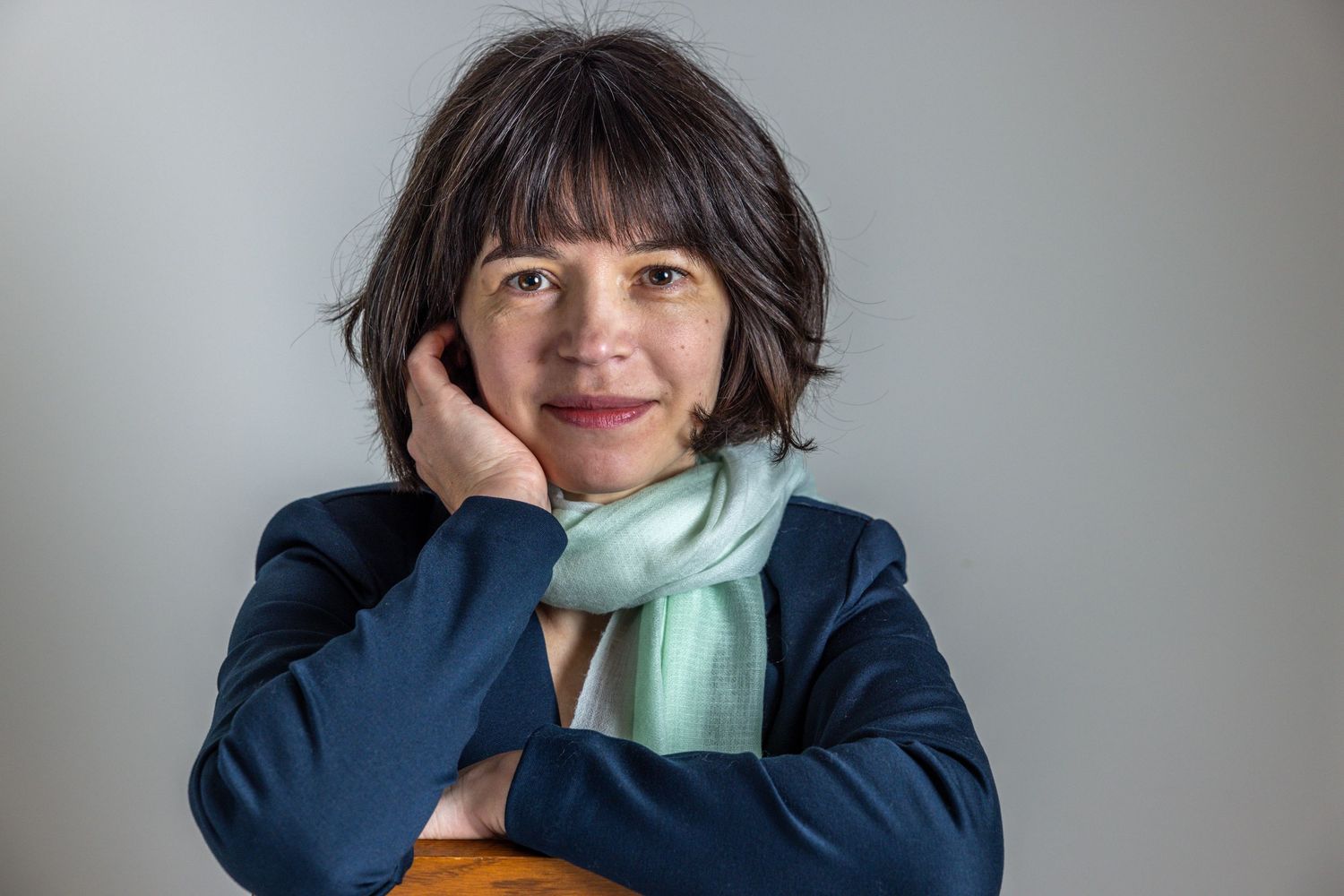 A professional in a navy blazer and mint scarf rests their chin on their hand while posing against a gray background.