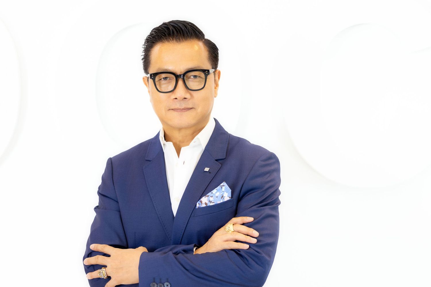 Business professional in navy blazer and glasses stands confidently with arms crossed against white background.