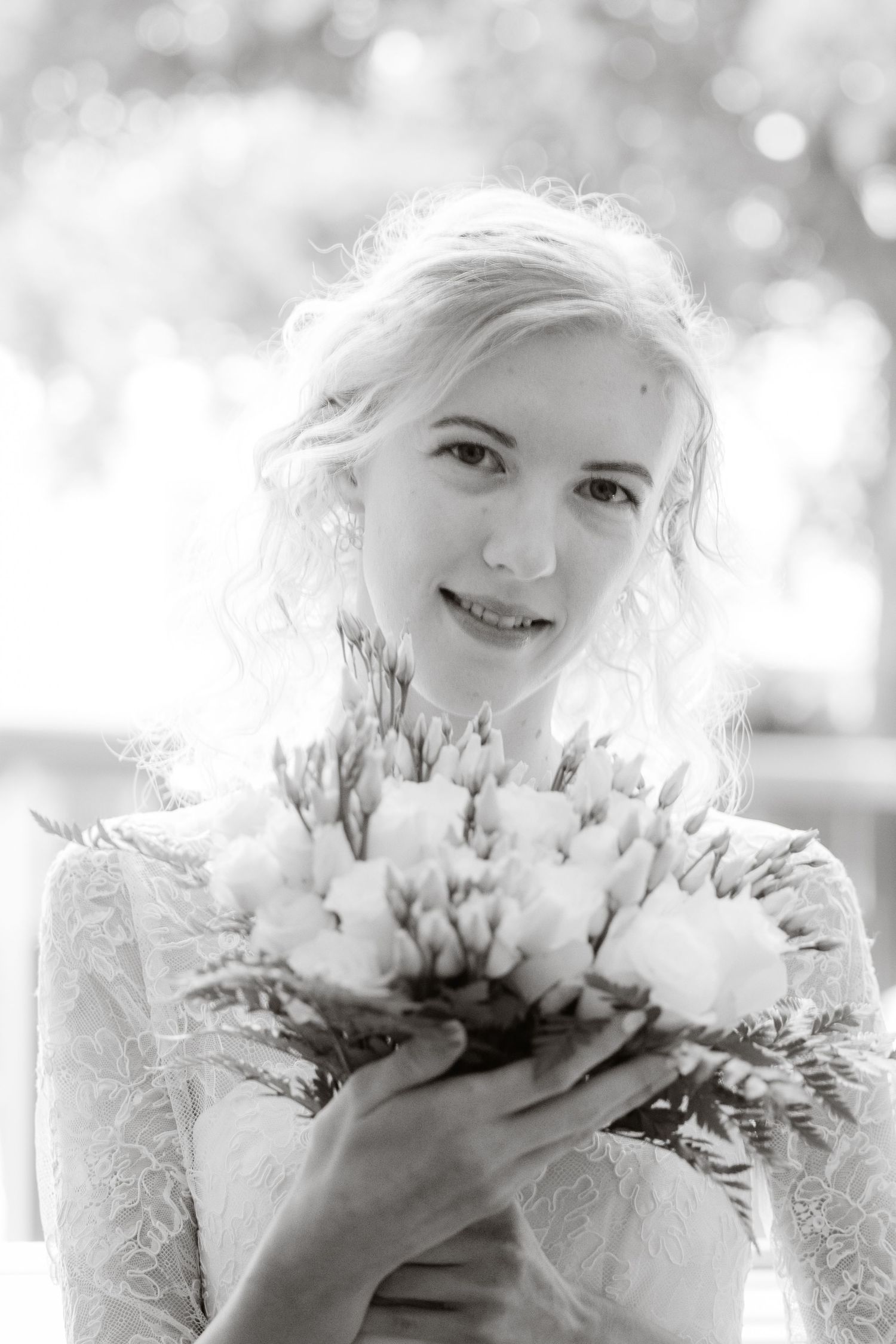 A bride in a lace gown holds a romantic bouquet of white flowers in this dreamy black and white portrait.