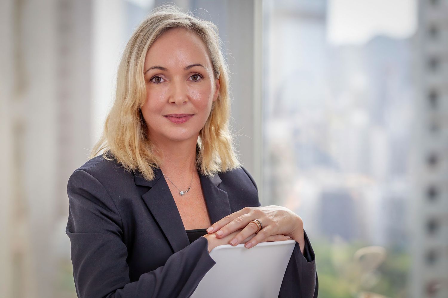 Professional businesswoman in black blazer poses confidently against urban backdrop with natural lighting.