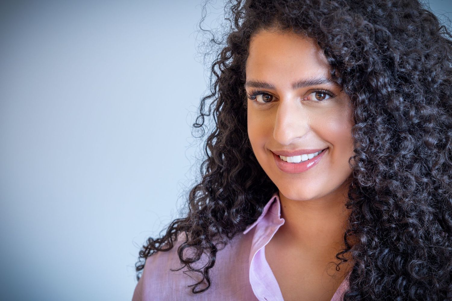 Professional headshot of someone with beautiful long curly dark hair wearing a lavender blouse against a gray background.