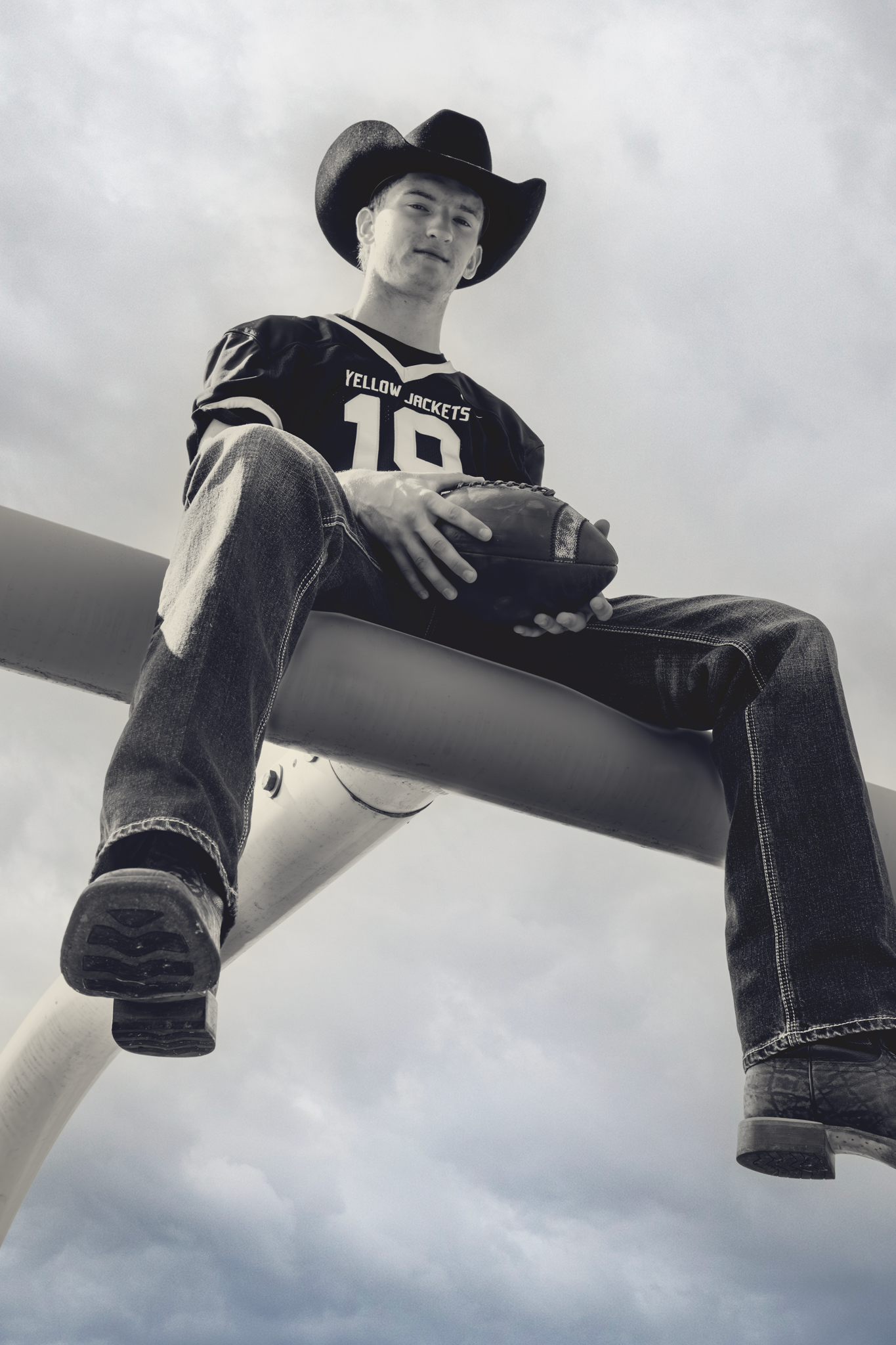 Black and white photo of person in cowboy hat and jeans sitting on a high ledge against cloudy sky.