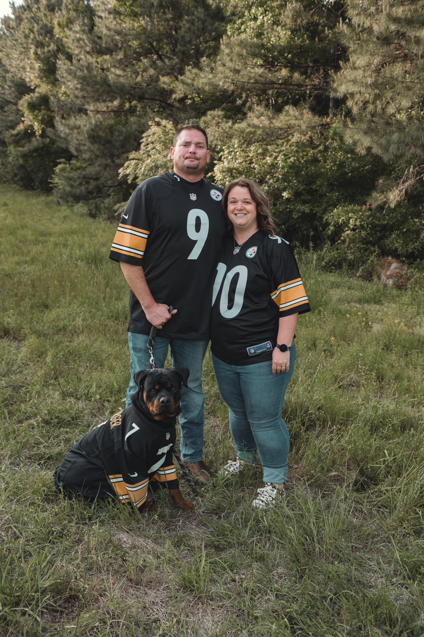 Couple in matching black and yellow football jerseys pose outdoors with their black dog wearing team gear.