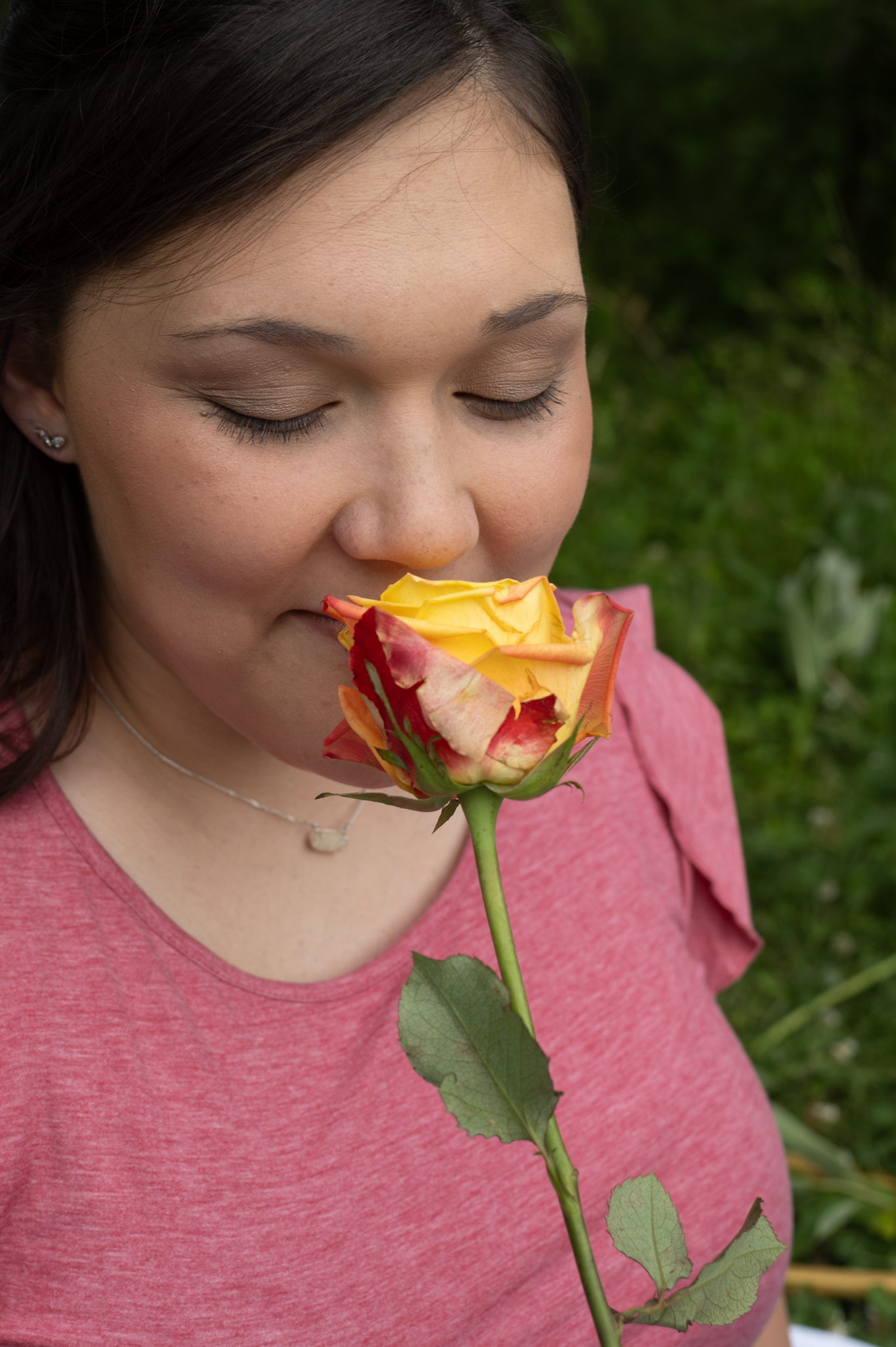 A person wearing a pink shirt smells a yellow and red rose in a garden setting.