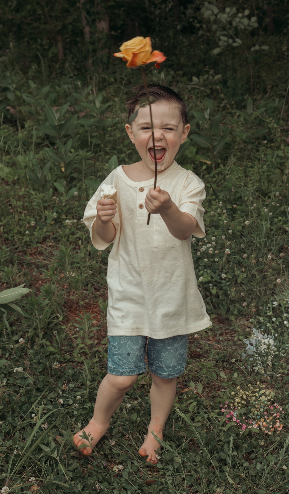 A joyful child in a white shirt and denim shorts stands in a garden holding an orange flower on a stem.