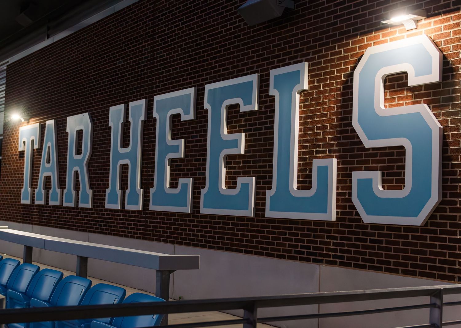 Illuminated Tar Heels signage mounted on a brick wall with stadium seating visible below.