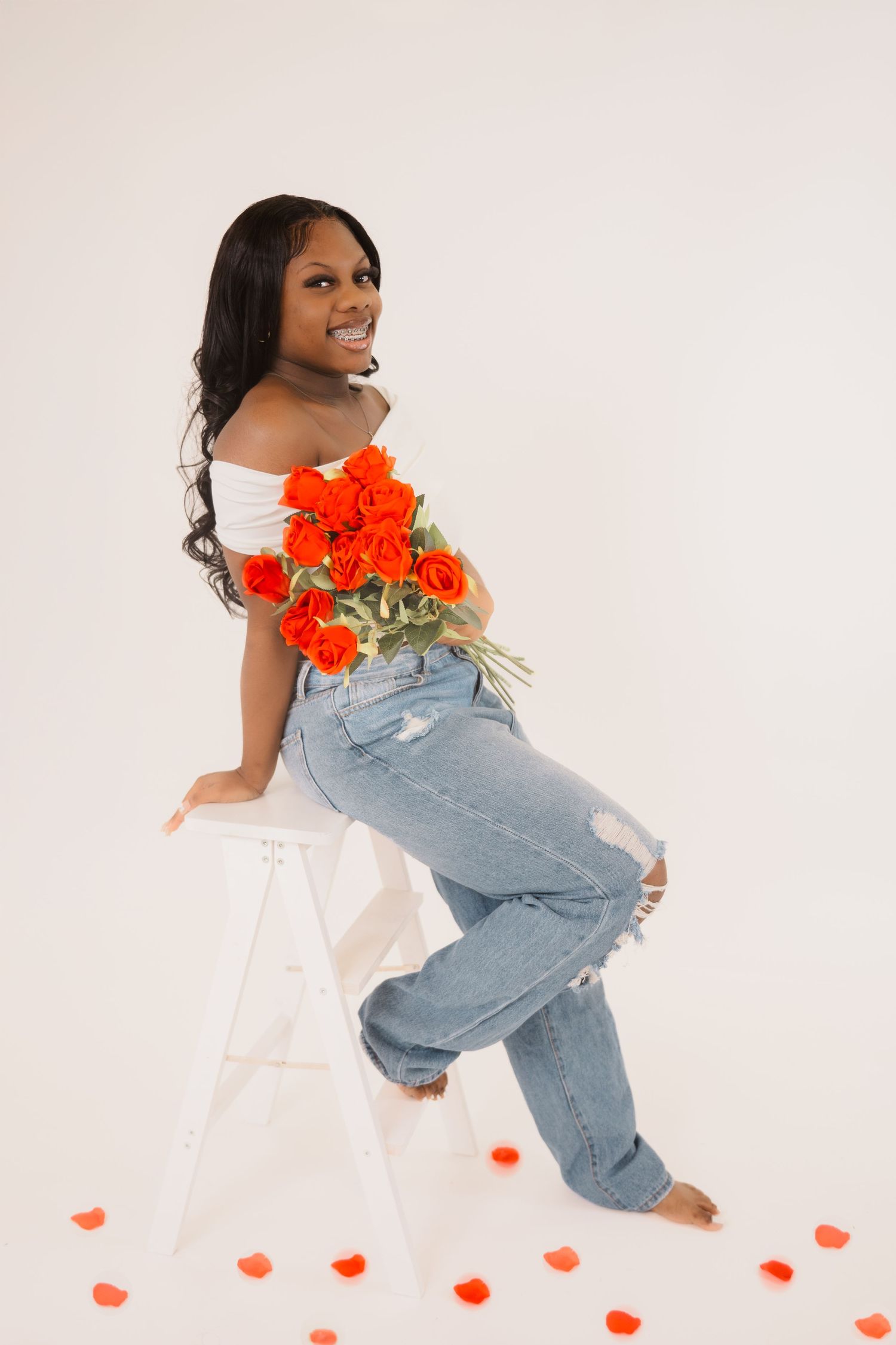 Happy woman in white off-shoulder top and jeans poses on stool with bright red flowers against light studio backdrop.