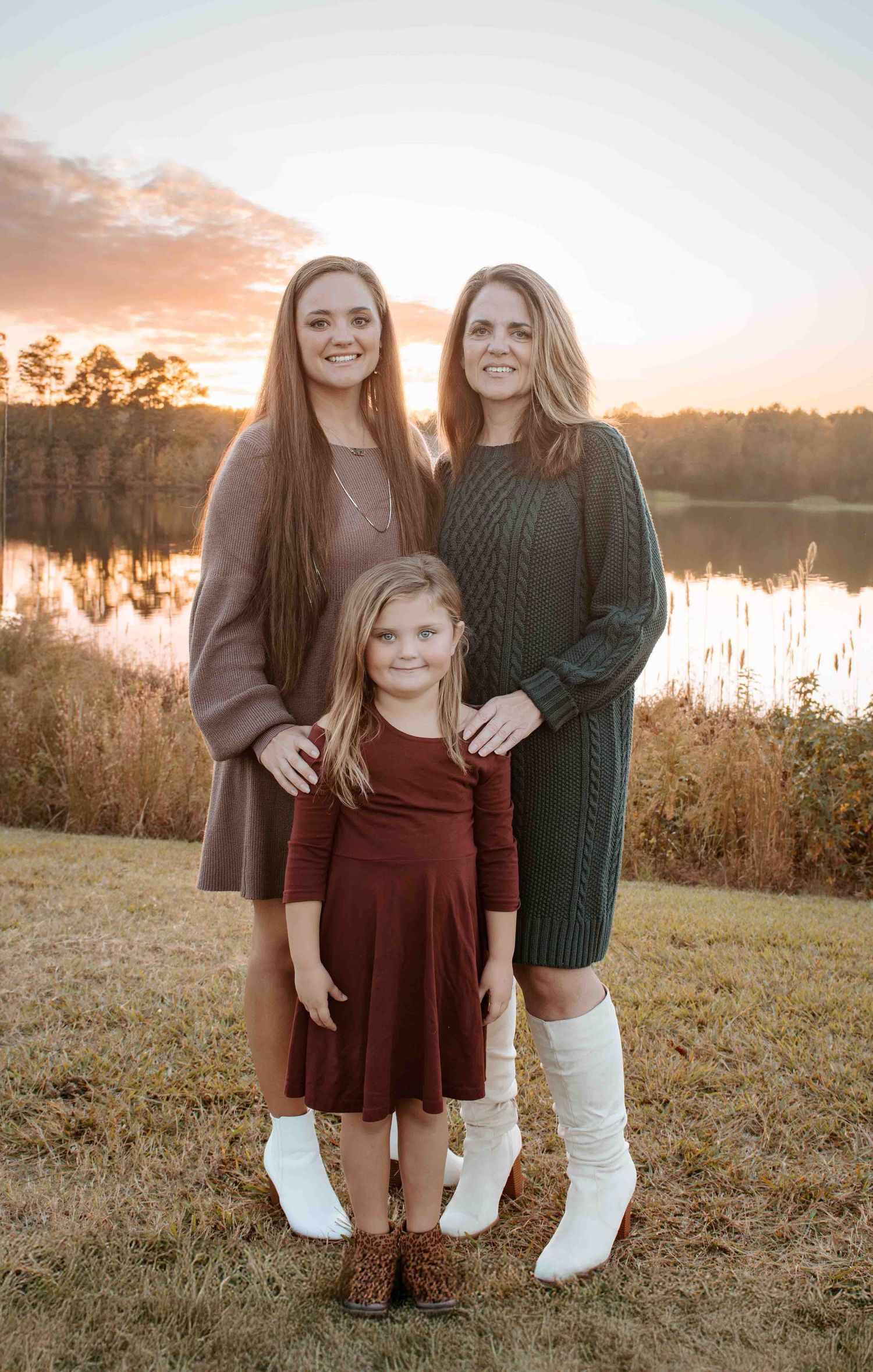 Three people stand together by a lake at sunset with trees in background wearing fall sweaters and dresses.