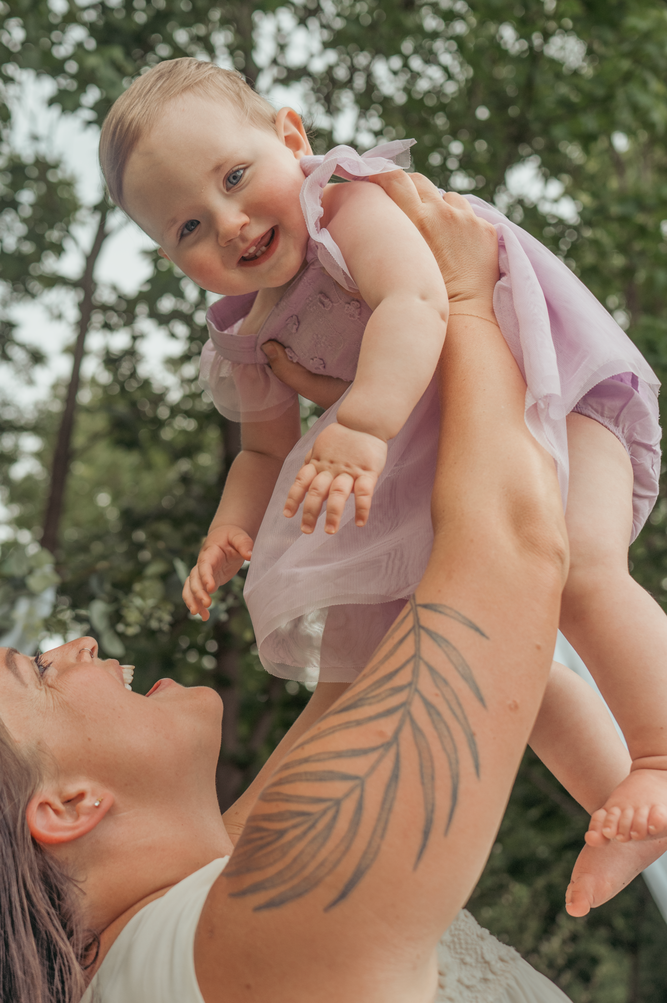 Joyful baby in pink dress being lifted up outdoors against green foliage by person with feather tattoo.