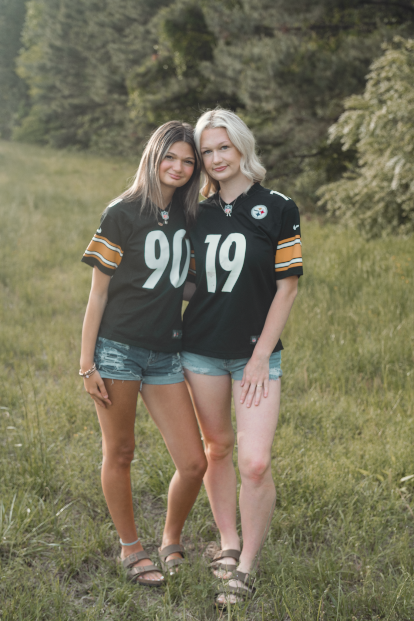 Two people wearing Pittsburgh Steelers jerseys with numbers 90 and 19 pose together in denim shorts on a grassy field.