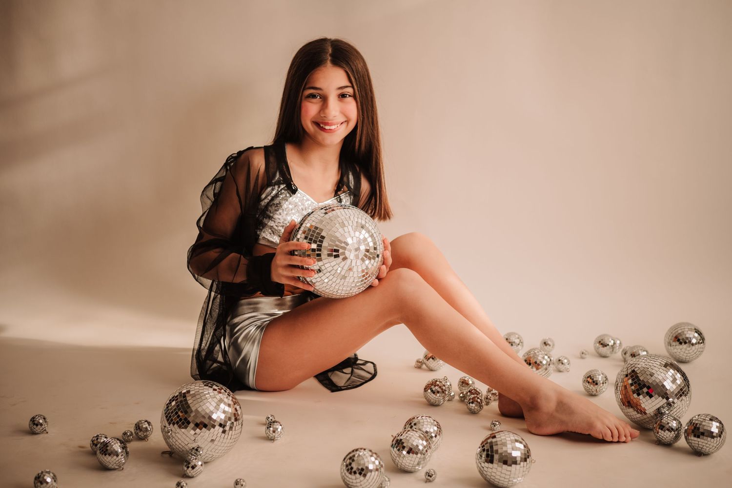 Studio photo with silver disco balls scattered on beige backdrop with figure seated among the shiny decorations.