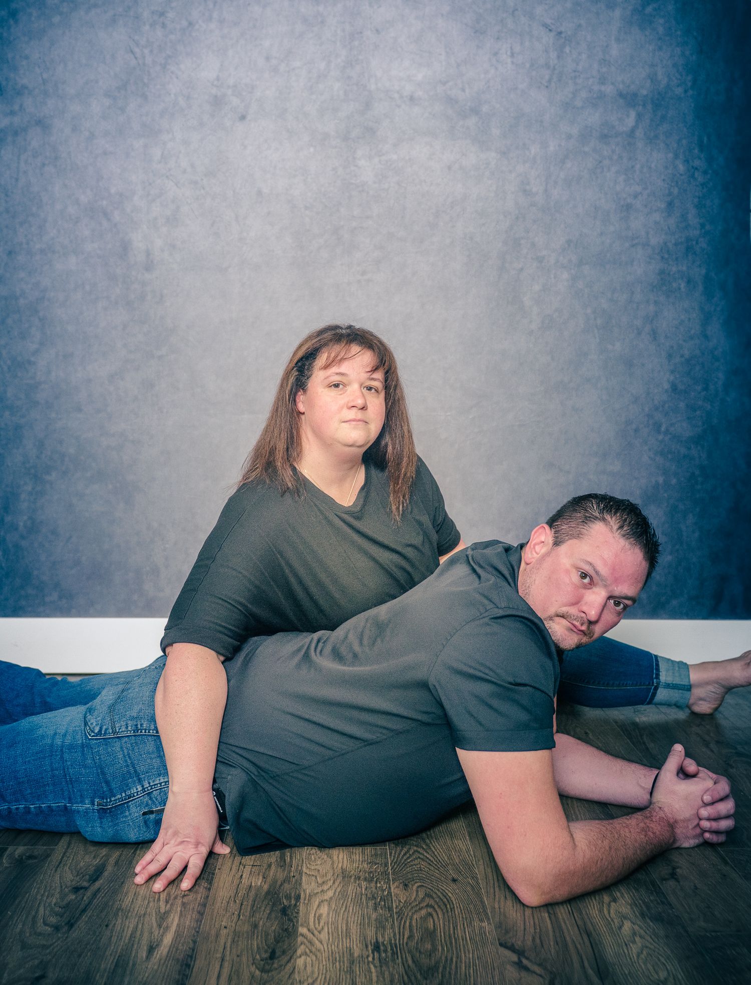 Two people in dark shirts and jeans pose together on a wooden floor for a casual portrait against a blue backdrop.