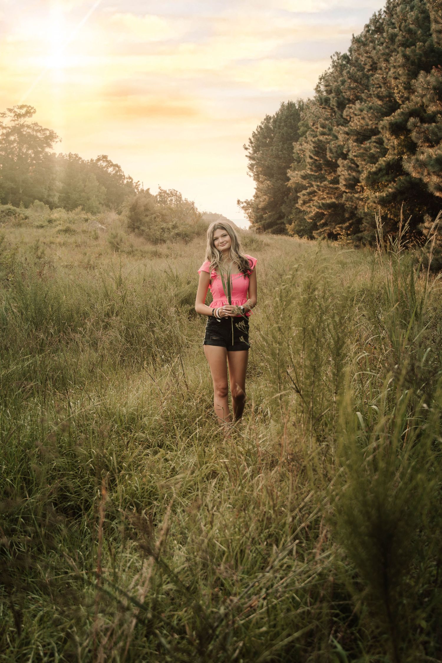 A dreamy sunset scene shows a person in a pink top and shorts standing in a tall grass meadow near trees.