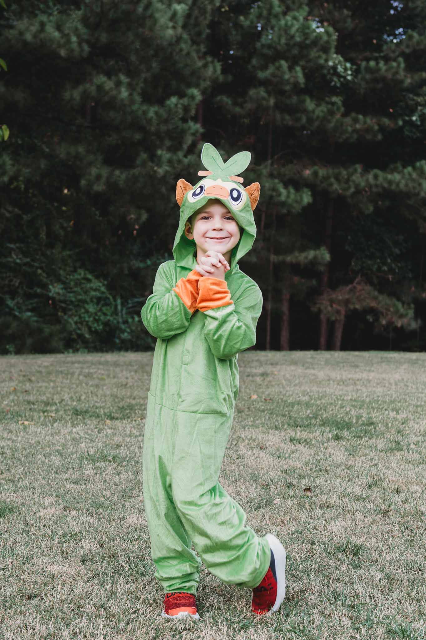 Someone wearing a cute green dragon or dinosaur costume onesie holds an orange drink while standing in a grassy yard.