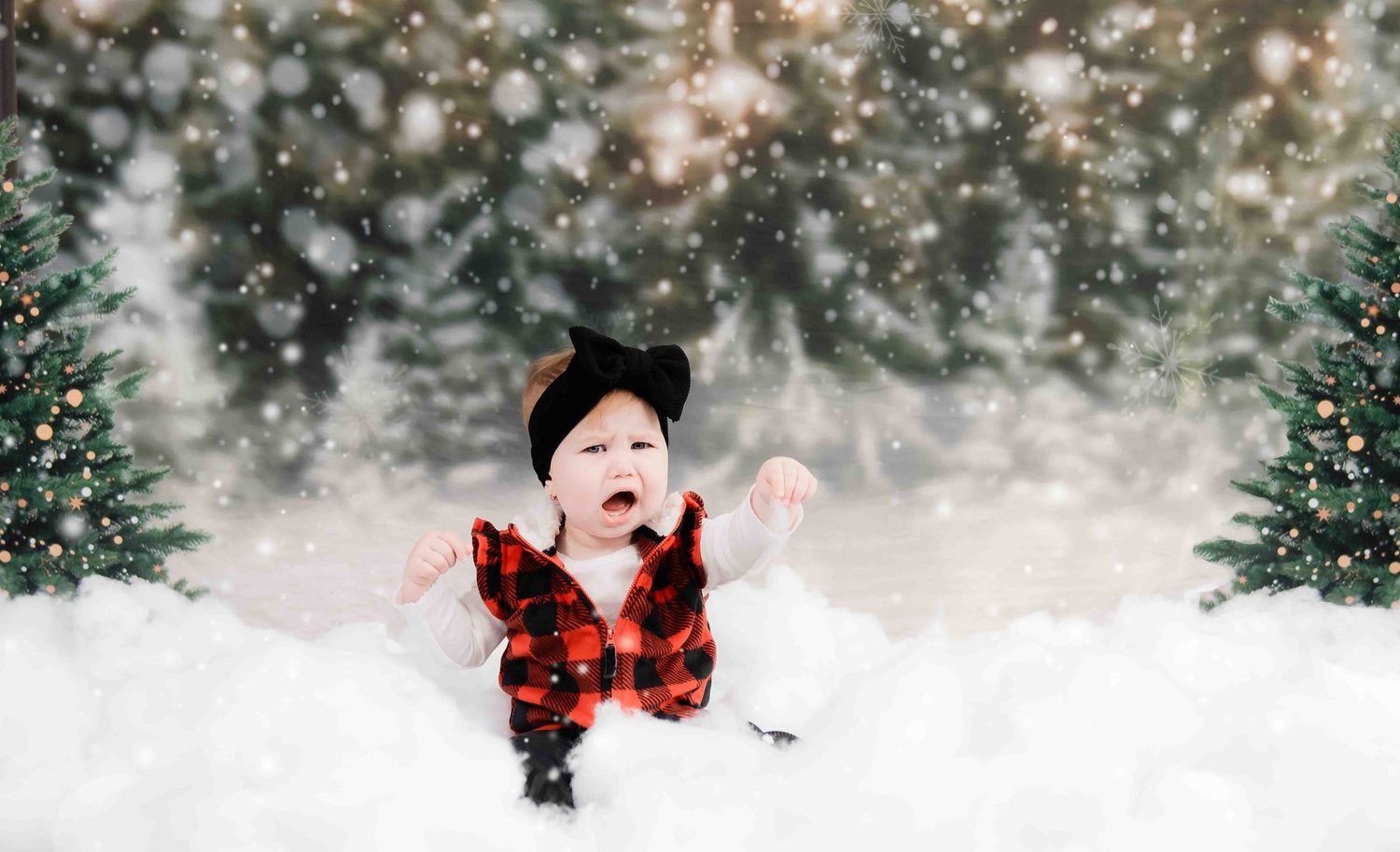A small child in red plaid vest plays in snowy scene with decorated Christmas trees in background.