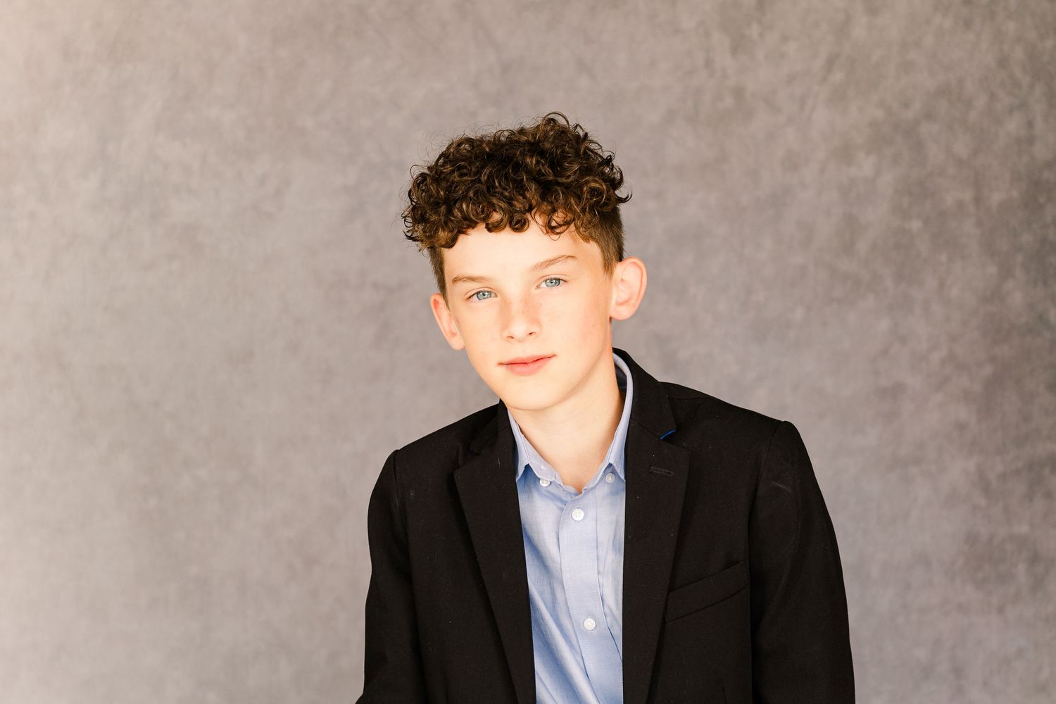 A teenager poses in a black blazer and light blue button-up shirt against a gray studio backdrop.