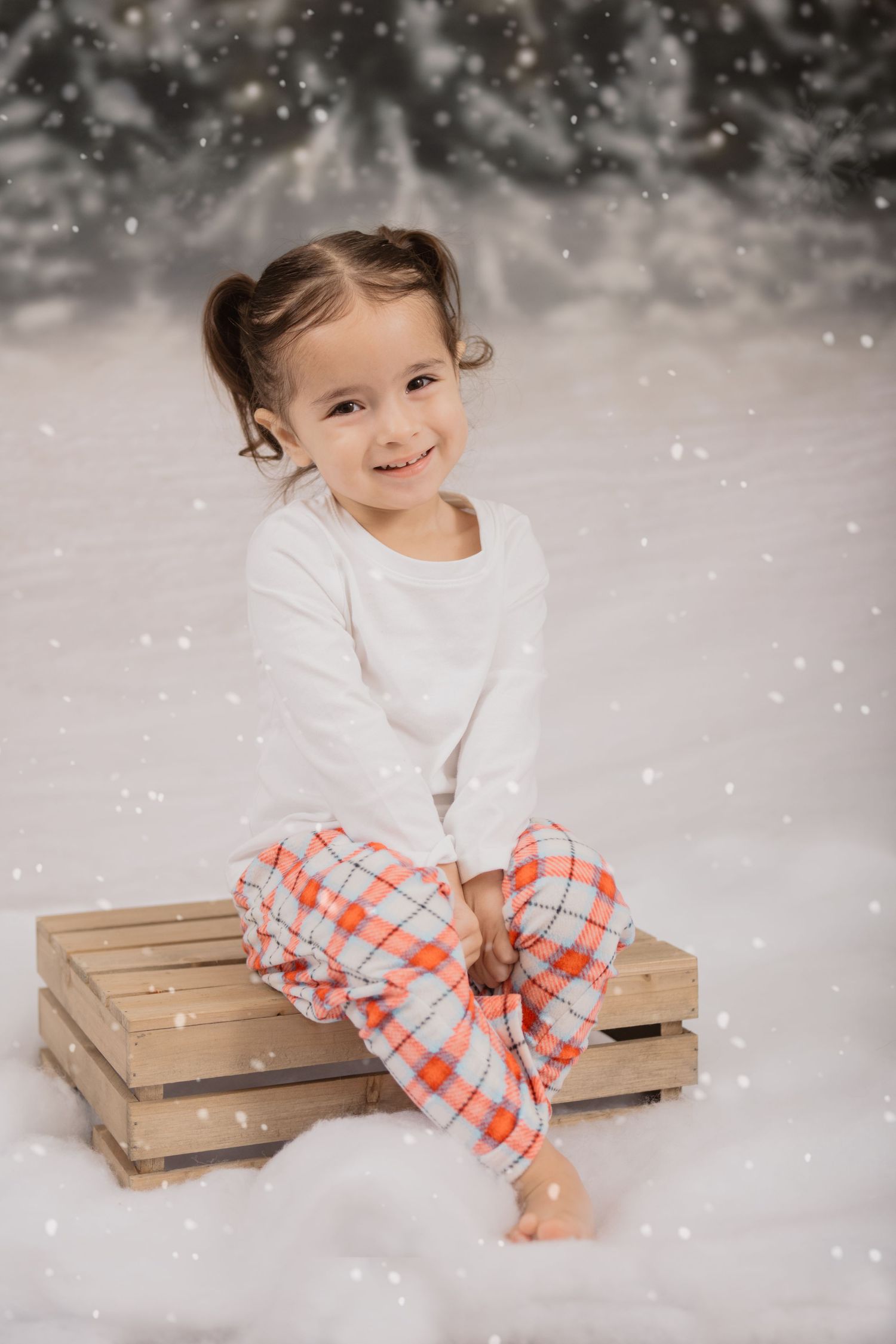 A young child in pajamas sits on a wooden crate during a snowy winter photo session.