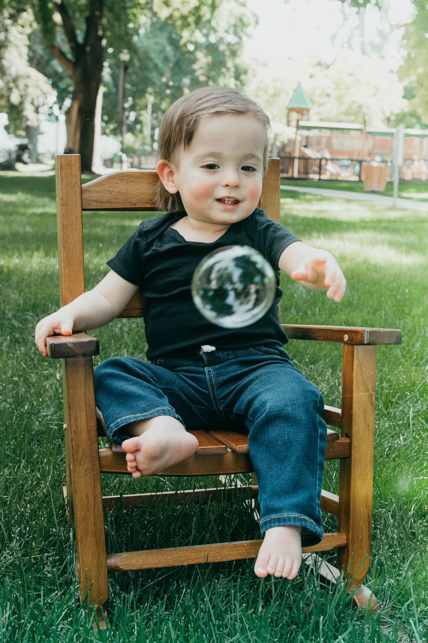 A small child sits in a wooden rocking chair outdoors, watching a soap bubble float by on a sunny day in the grass.