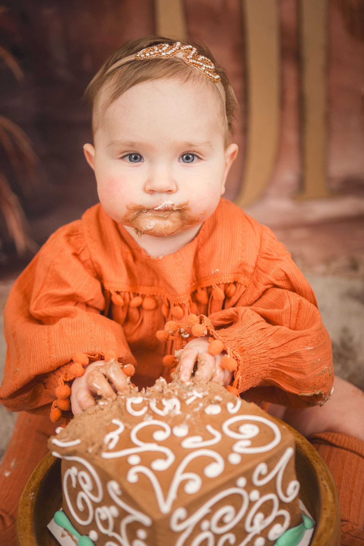 A beautifully decorated brown cake sits in front of a small child wearing a bright orange dress.
