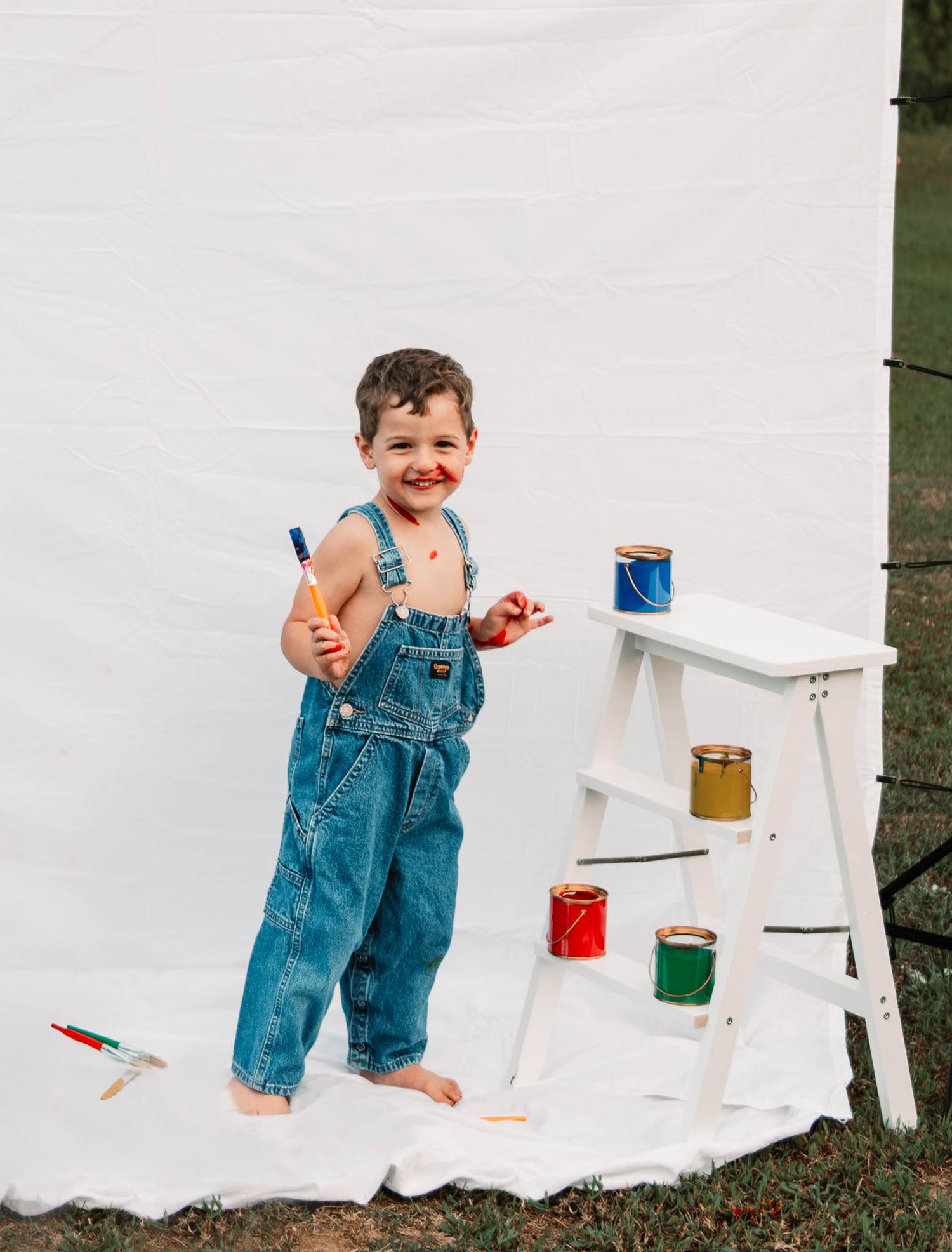 Small child in denim overalls stands barefoot next to white stepladder with colorful paint jars and paintbrushes.