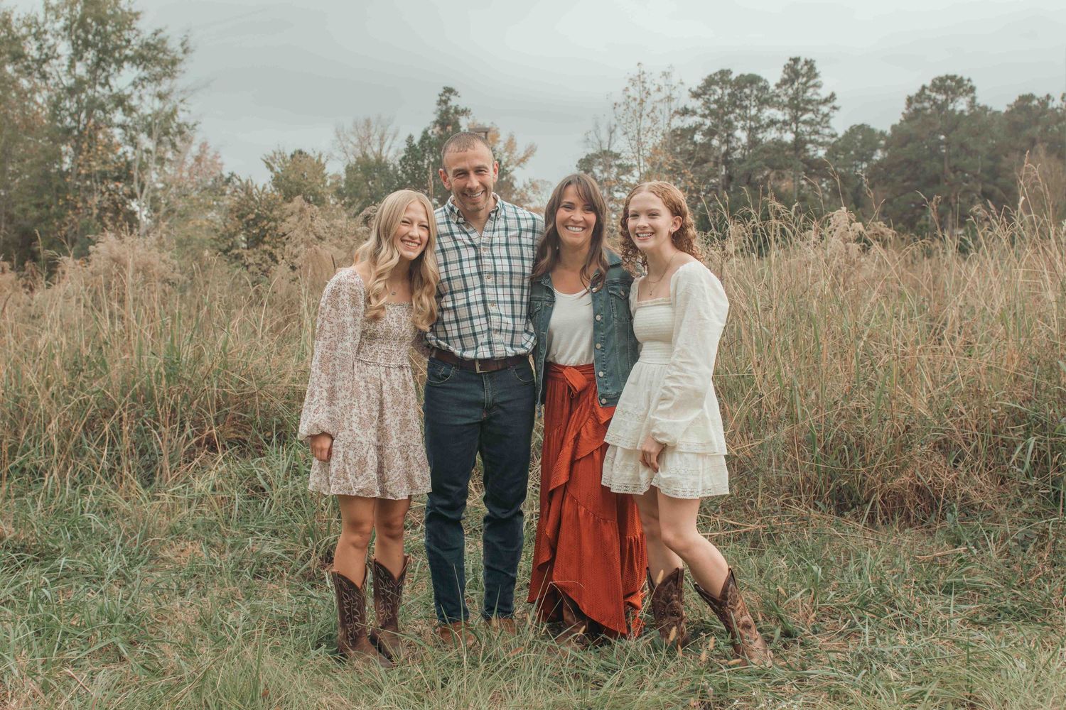 A group of four people stands together in a rustic field with tall brown grass and trees in the background.