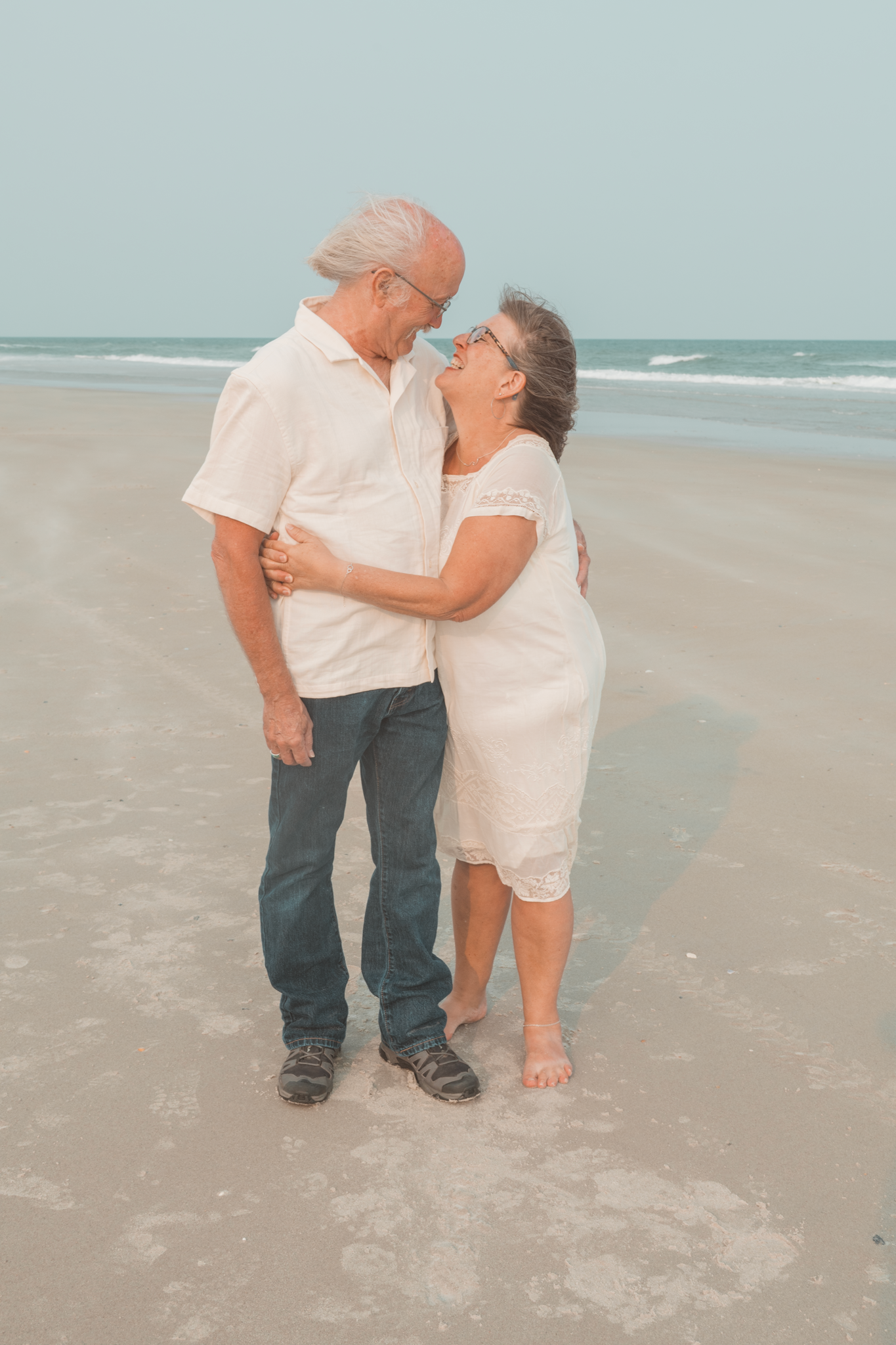 An elderly couple in white clothing embraces lovingly on a peaceful beach with ocean waves in the background.