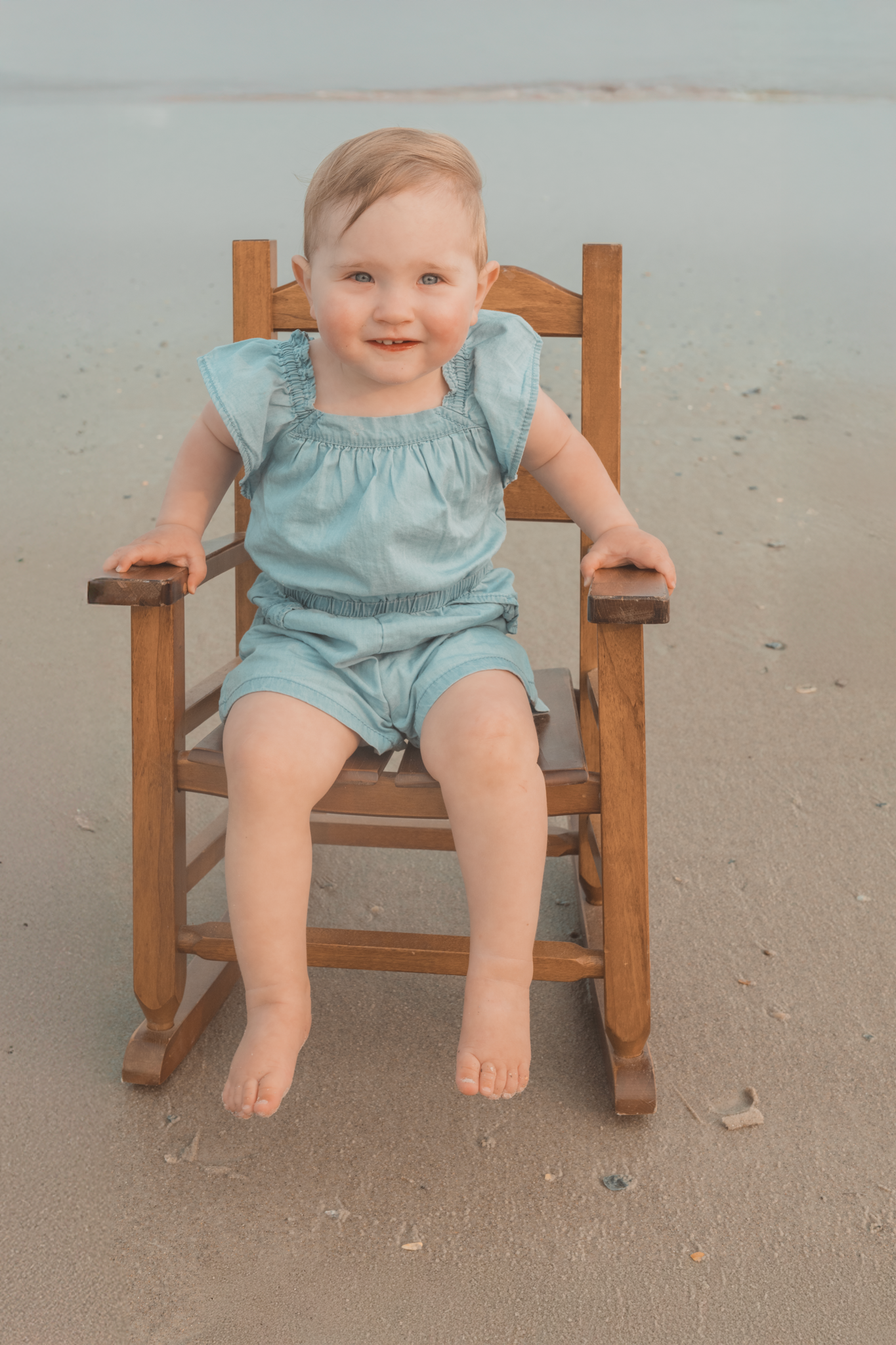 A child wearing a light blue outfit sits smiling in a wooden rocking chair on the beach.