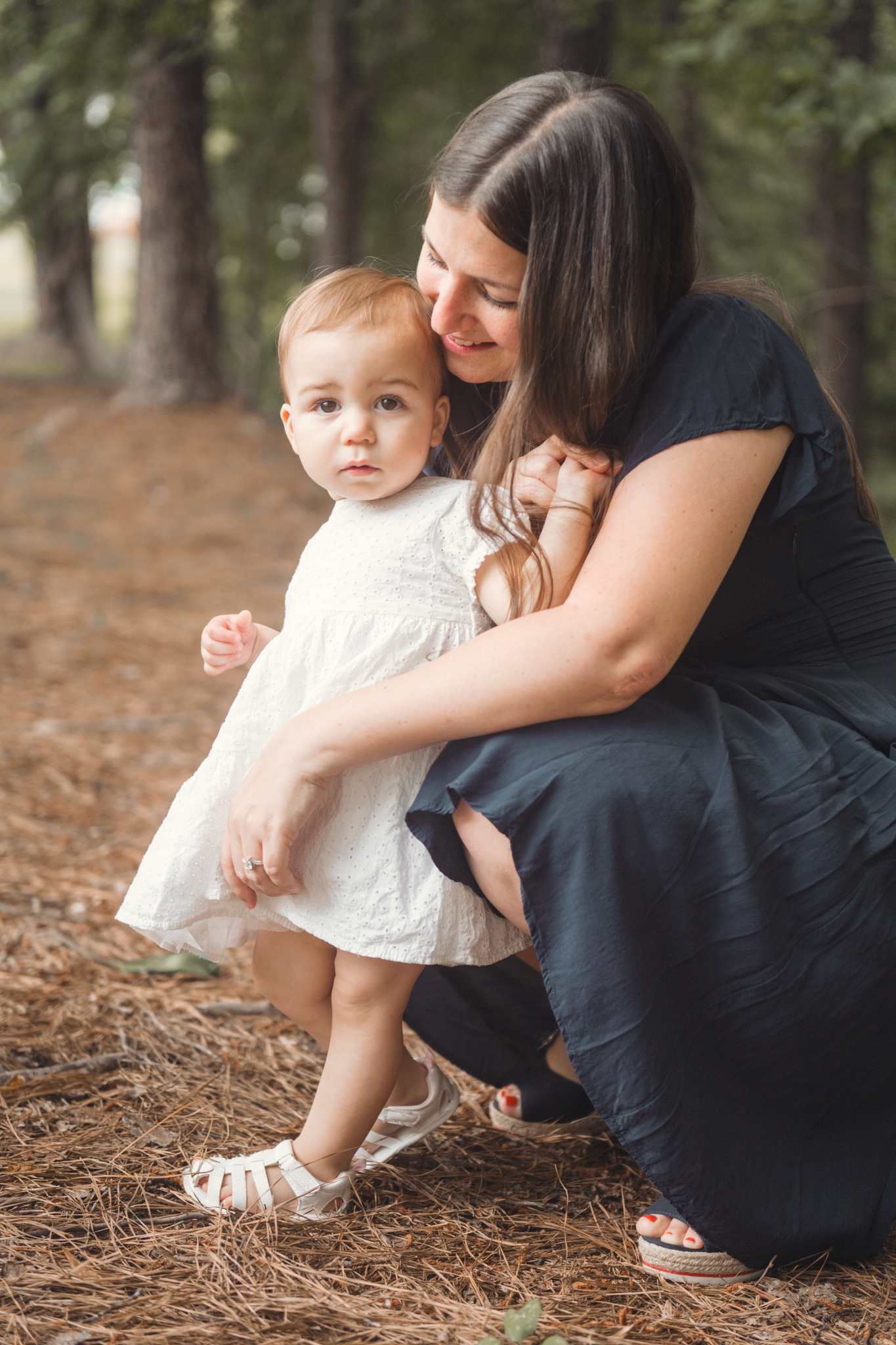 A heartwarming outdoor portrait of a caregiver in a navy dress kneeling beside a baby wearing a white lace dress.