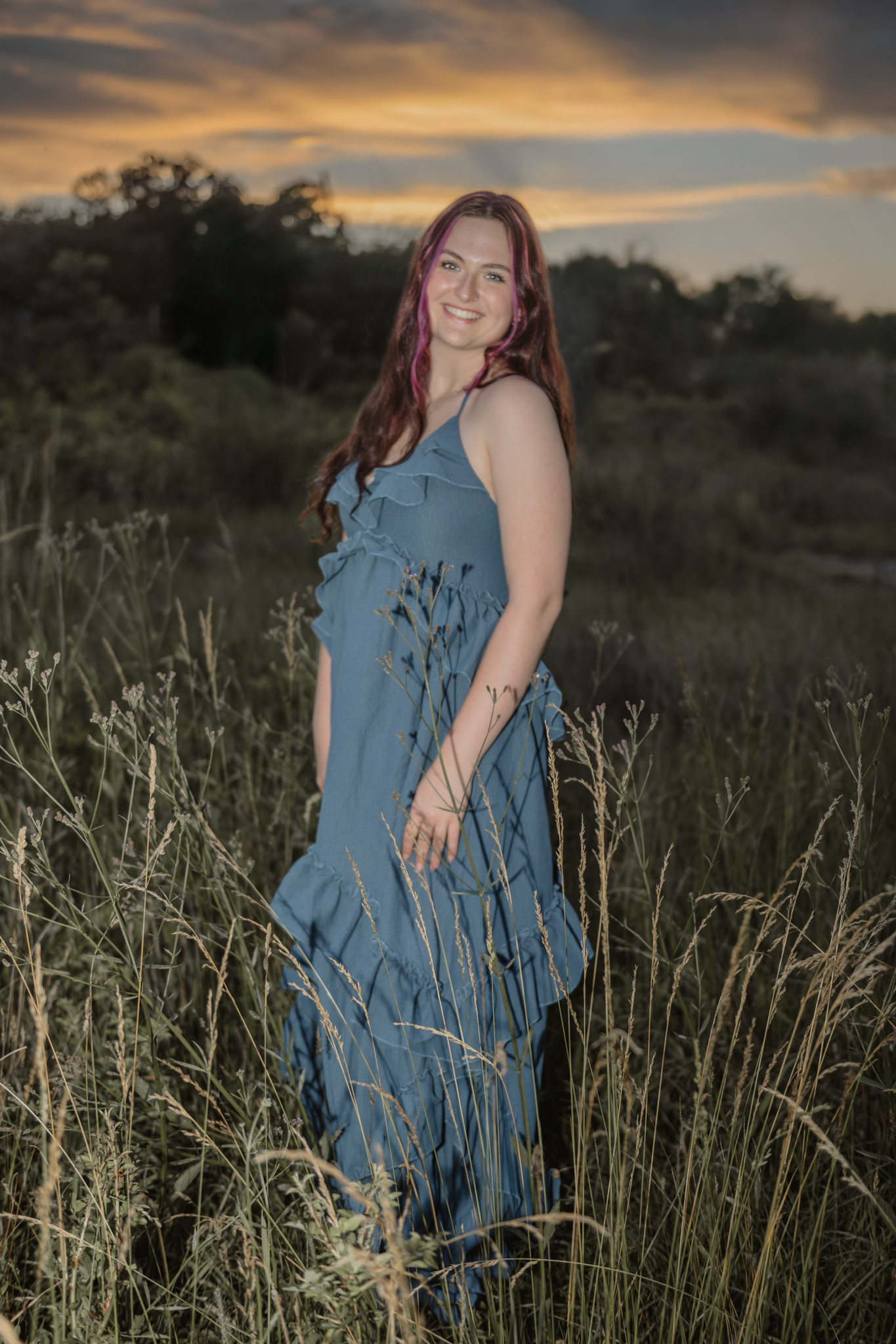 Haylee in a flowing blue dress stands in tall grass during a golden sunset with dramatic clouds in the background during her senior session.