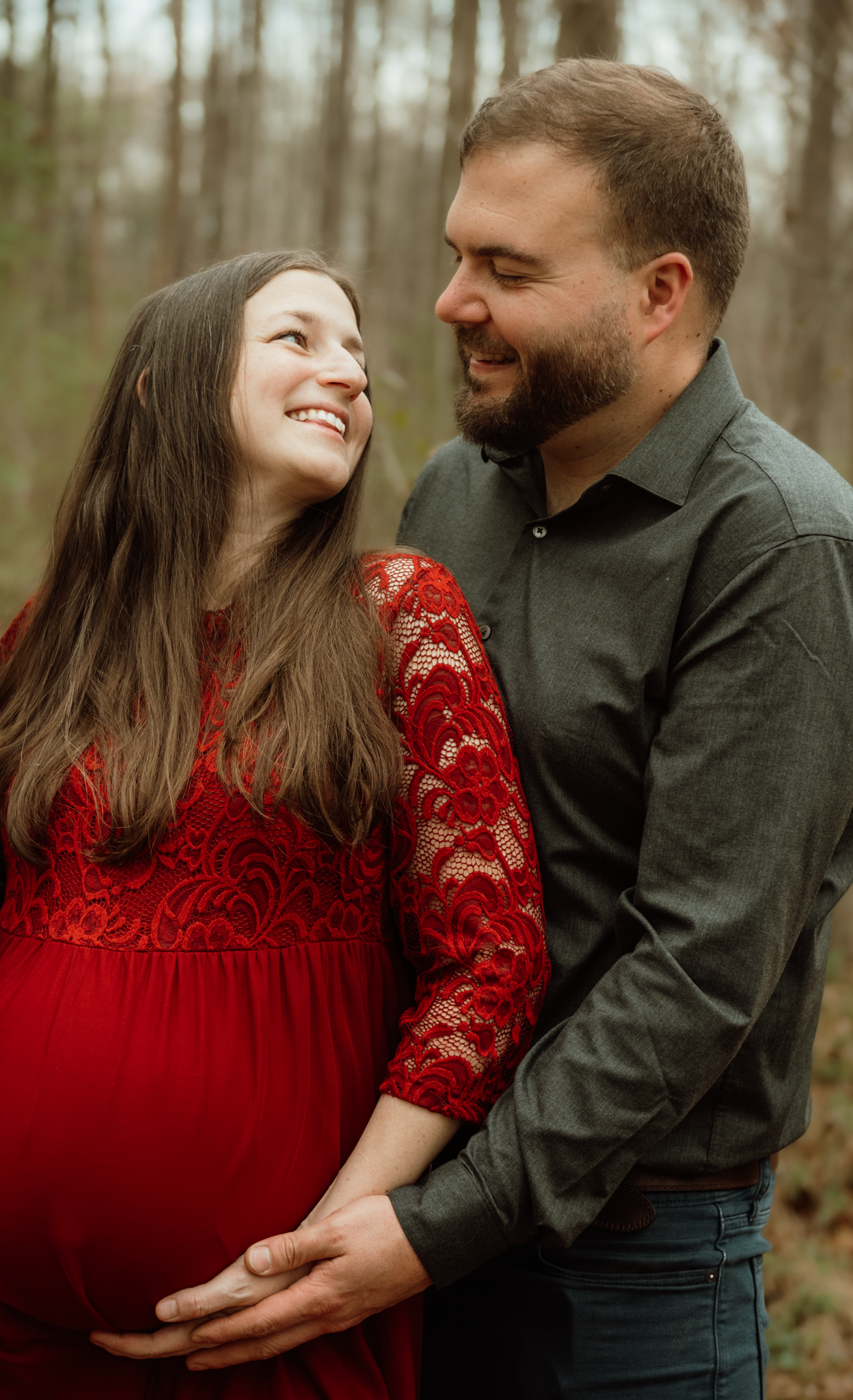 A smiling pregnant woman in a red lace dress stands with her partner outdoors in a wooded setting for a maternity photo.