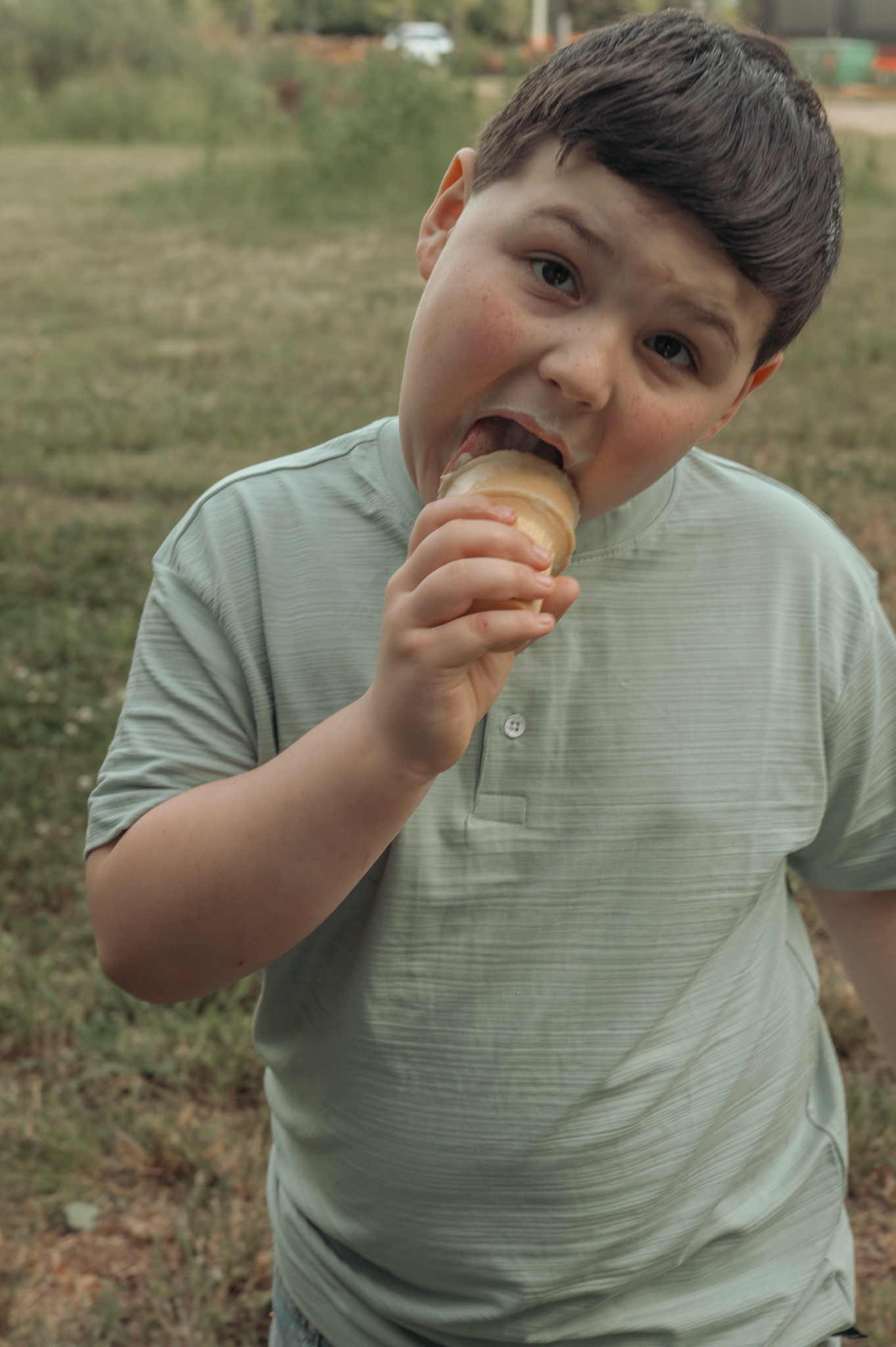 Child in a light colored polo shirt eating an ice cream cone outdoors in a grassy field.