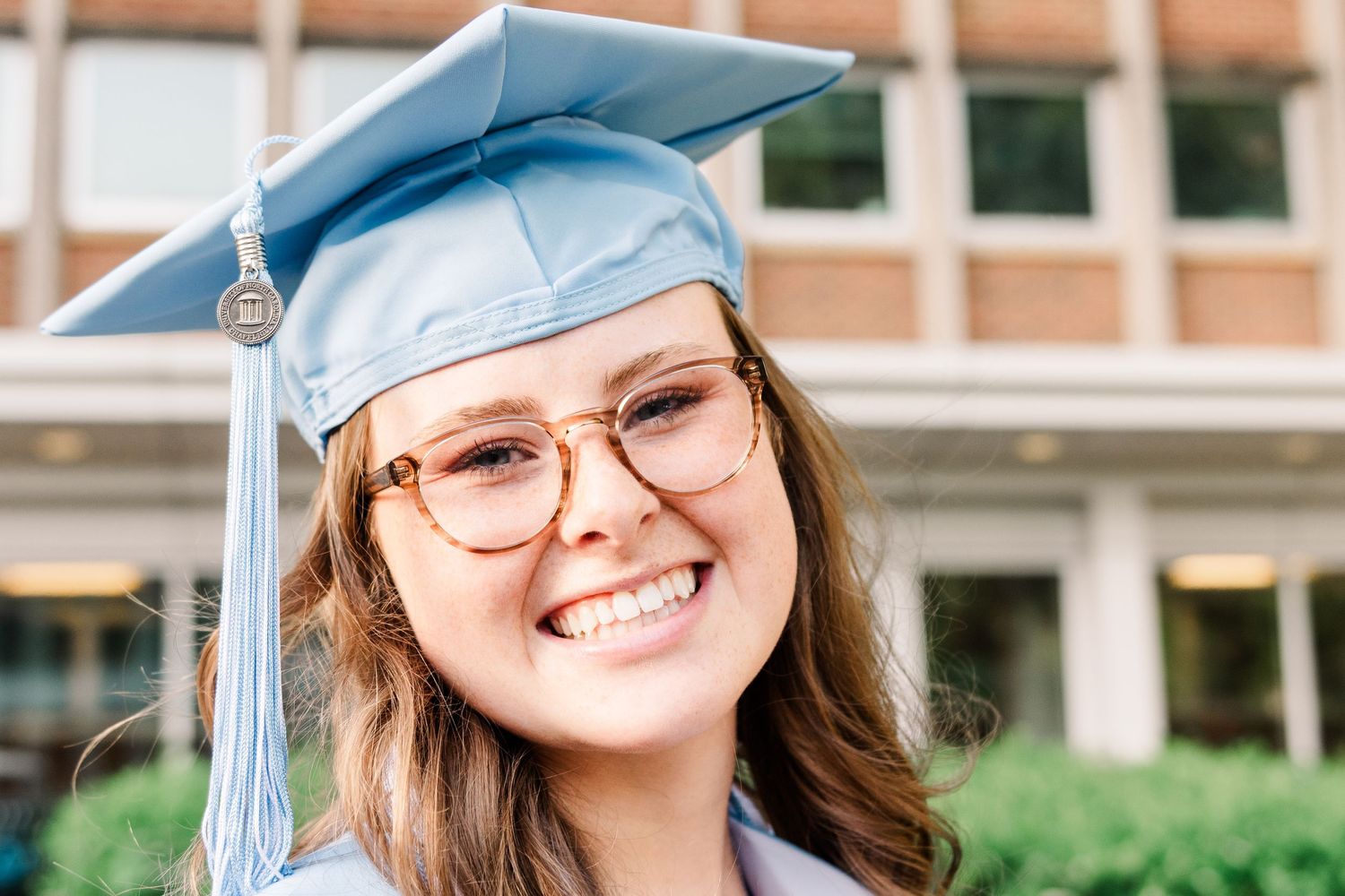 Graduate wearing light blue cap and gown with tassel smiles brightly while wearing glasses on graduation day.
