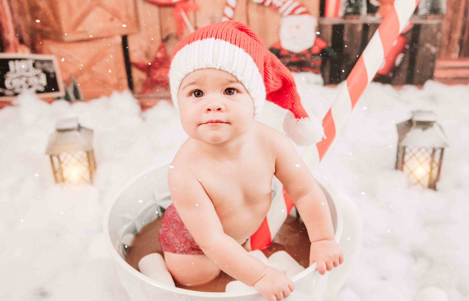 A small child wearing a pink Santa hat and red diaper sits on white plush fabric with holiday lanterns in the background.