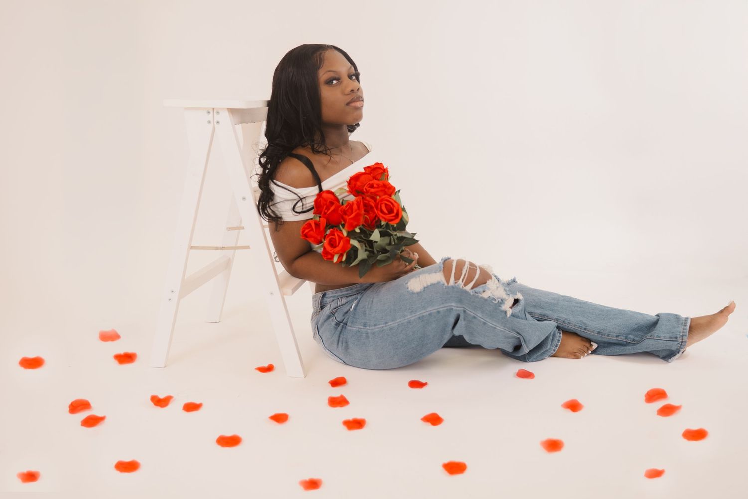 Model reclines on white backdrop with red roses and scattered petals, wearing distressed jeans and off-shoulder top.