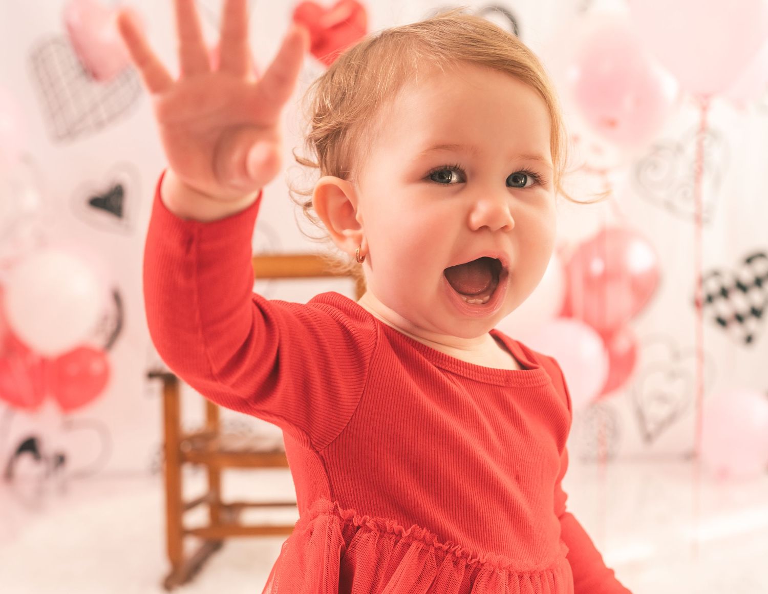 Small child wearing red dress waves excitedly against pink and white background with hand prints on wall.